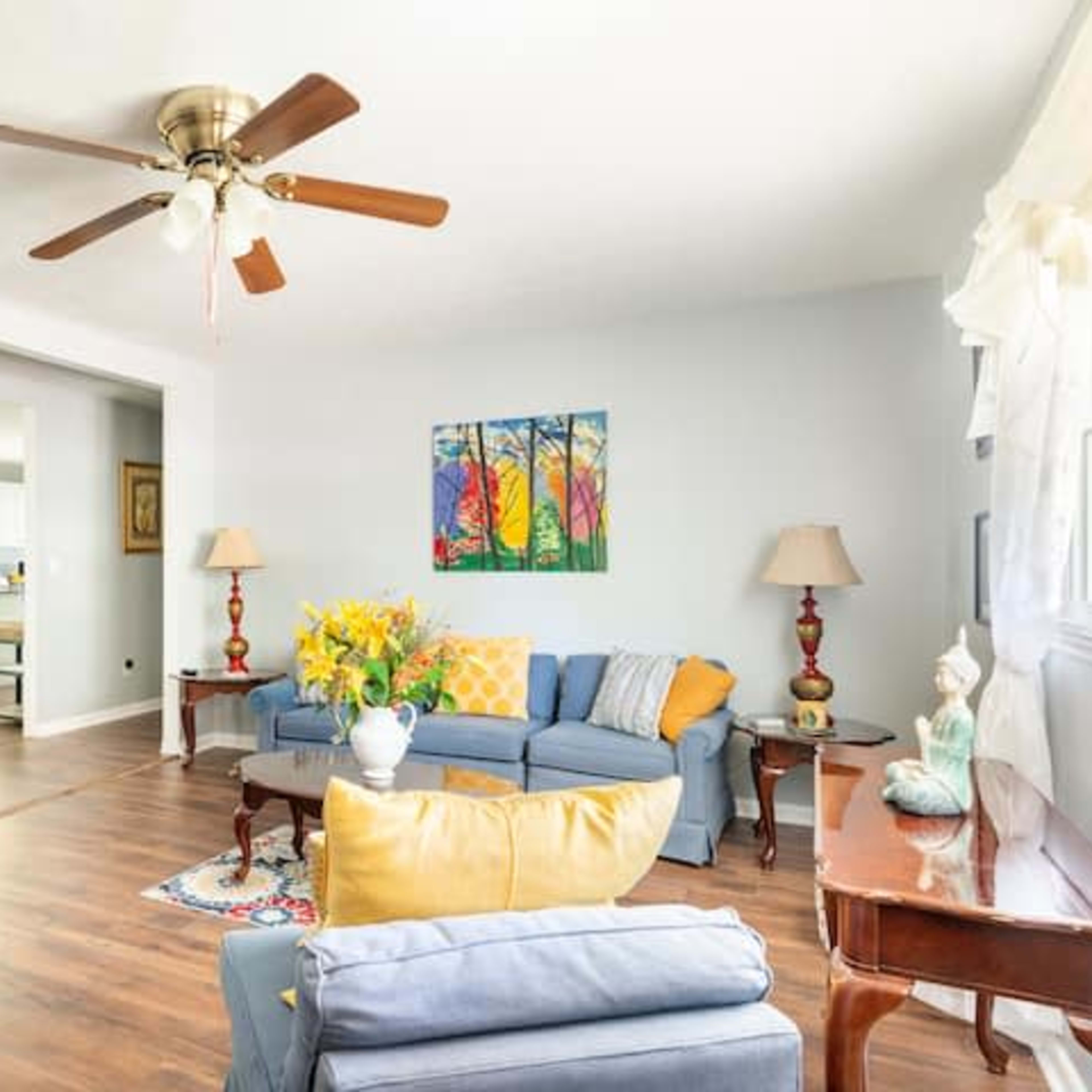 A well-lit living room features blue furniture, a ceiling fan, and colorful wall art, with a view into a connected kitchen area.