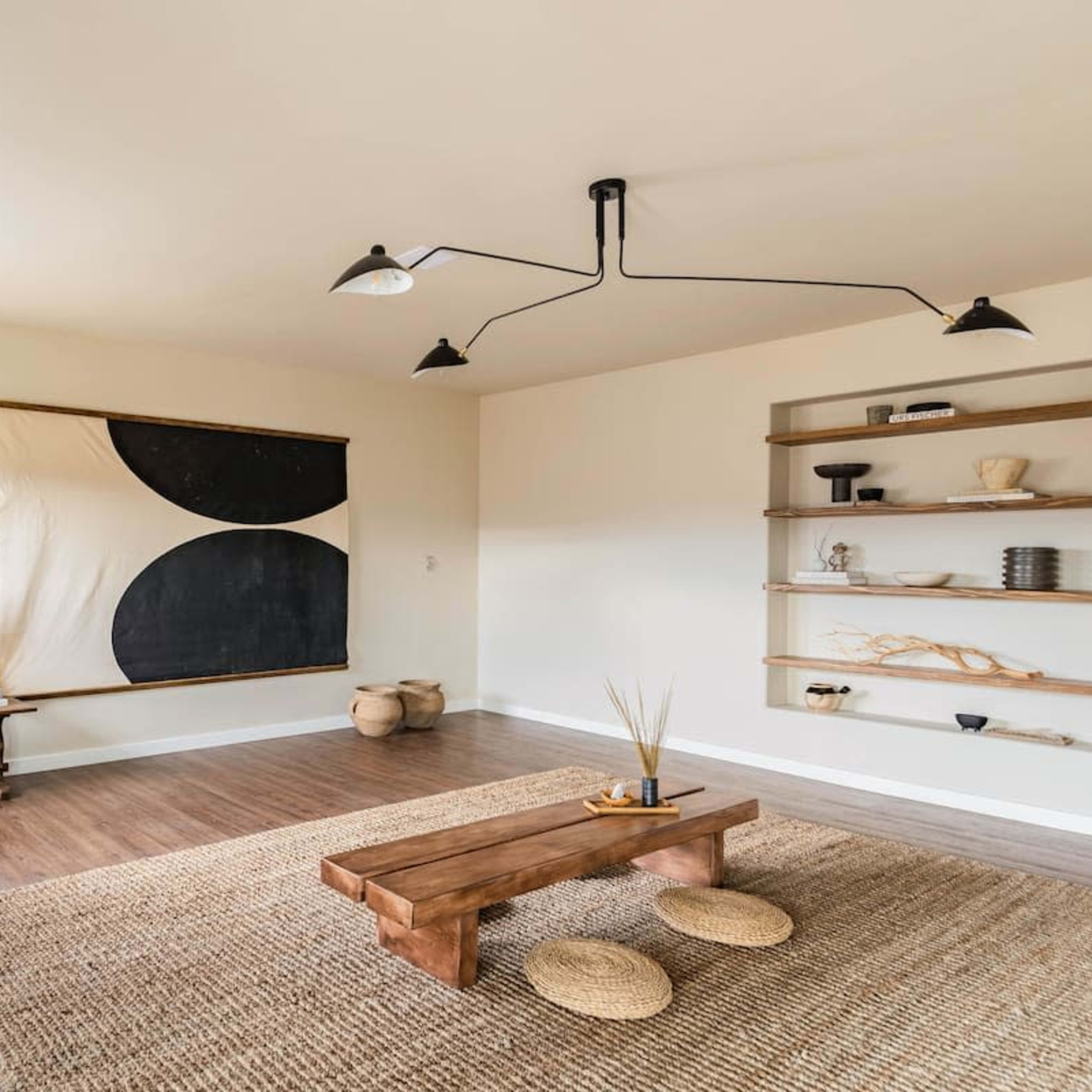 A minimalist living room features a low wooden coffee table, a woven rug, a wall art piece with black and beige shapes, and wooden shelves displaying decorative items.