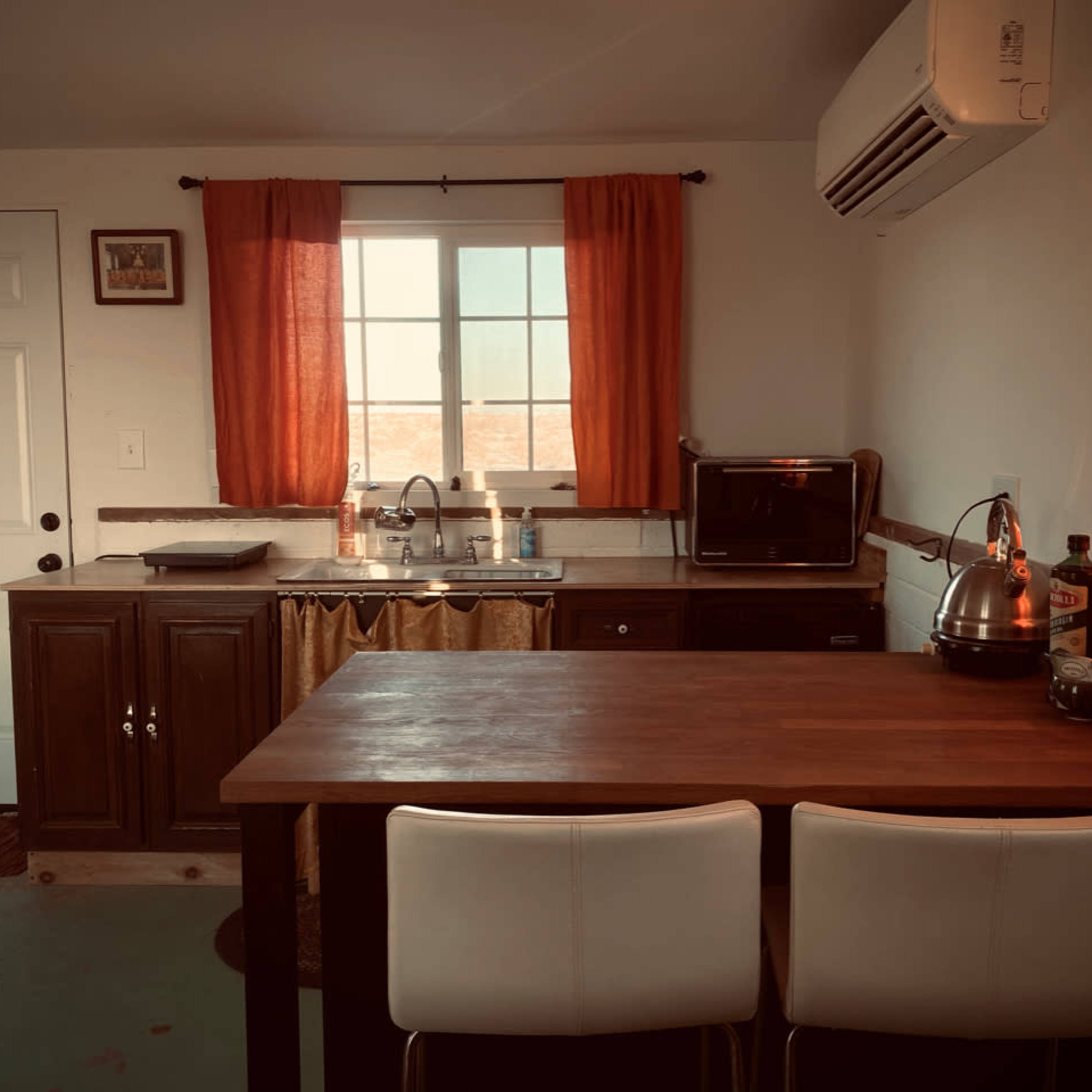 A small kitchen with a sink, microwave, and a wooden table with two white chairs, complemented by orange curtains at the window.