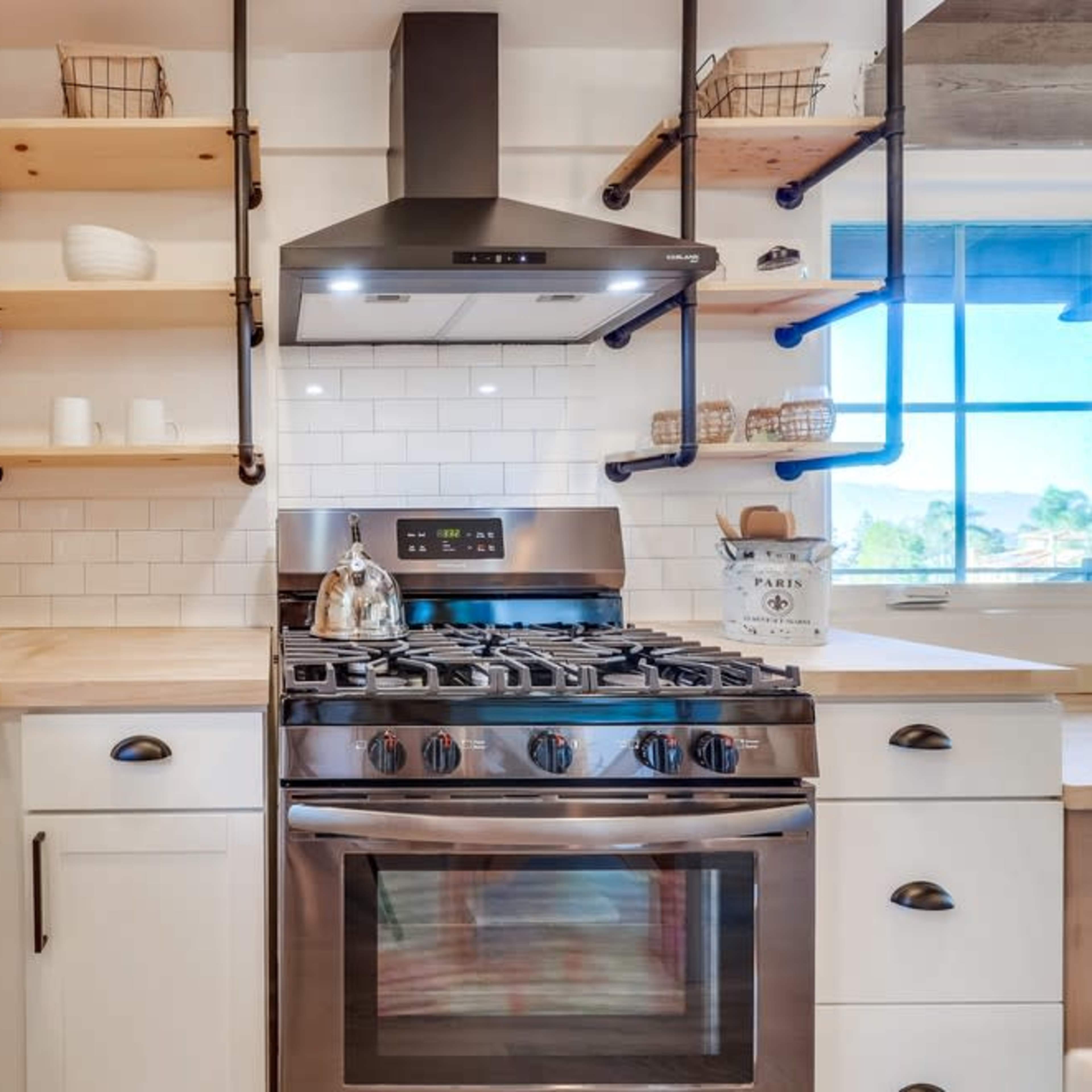 A modern kitchen features wooden shelves, a gas stove, and a stainless steel hood above a countertop.