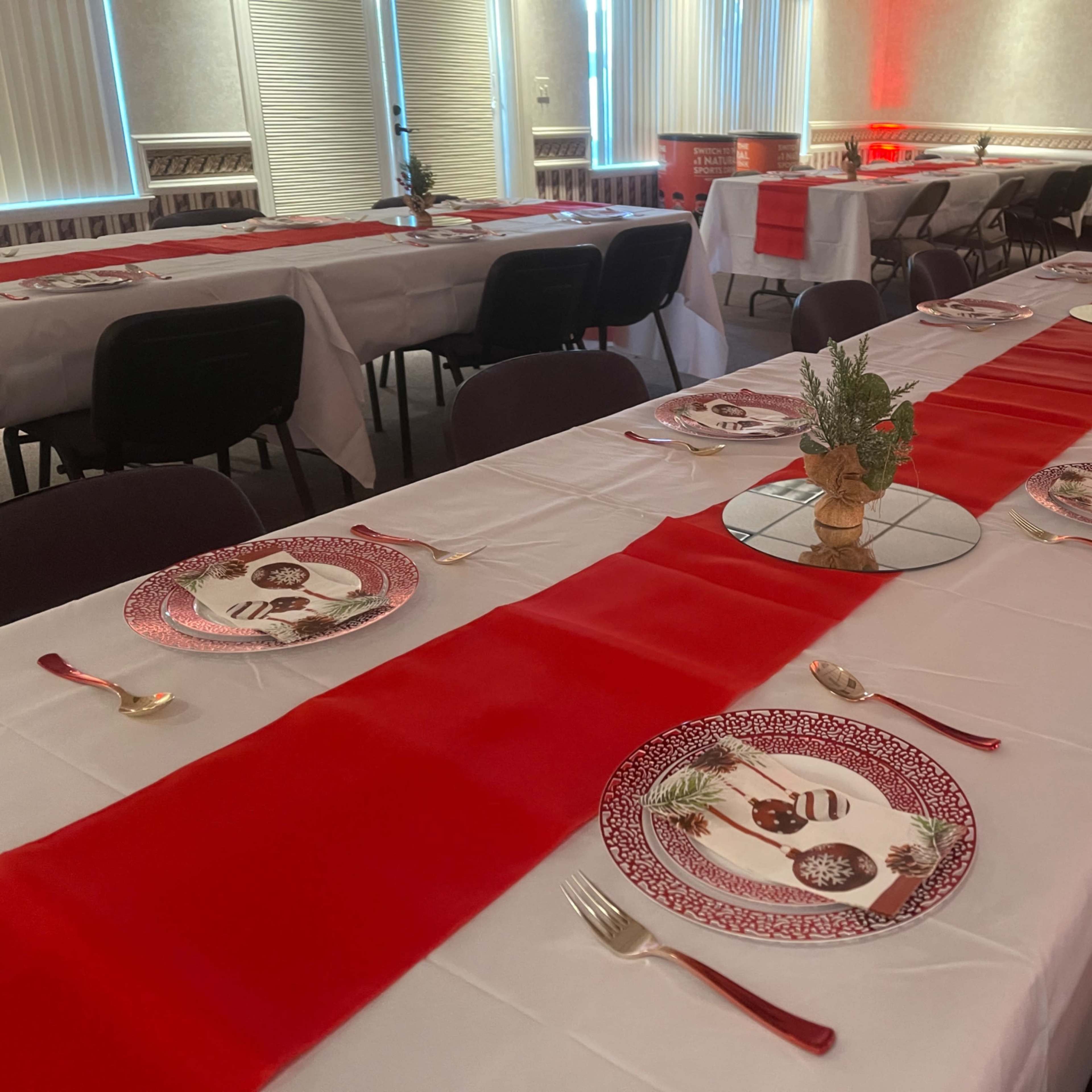 A dining setup features multiple tables covered with white tablecloths and red runners, arranged with decorative plates and utensils.