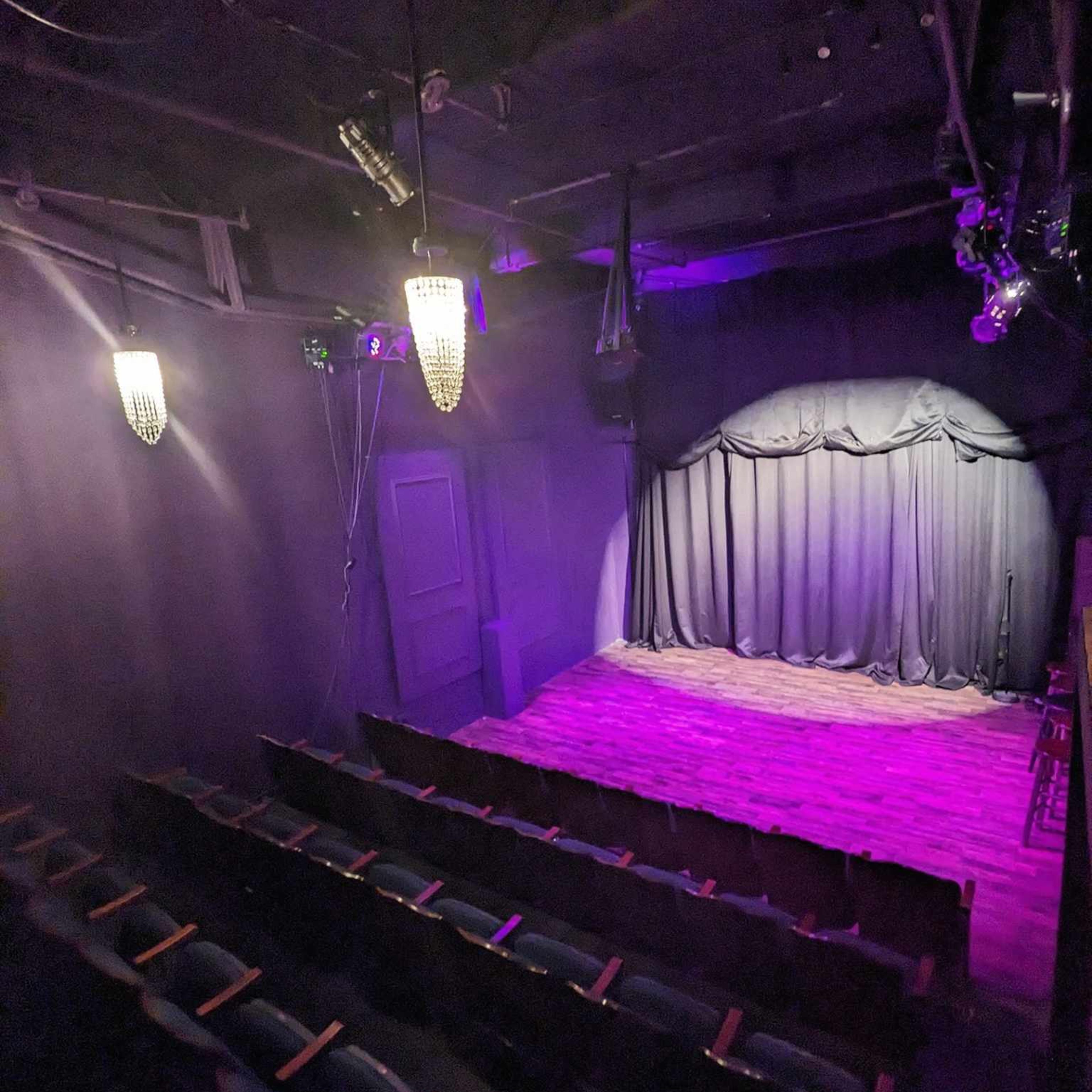A dimly lit theater with empty seats facing a stage covered by black curtains and illuminated by purple lights.
