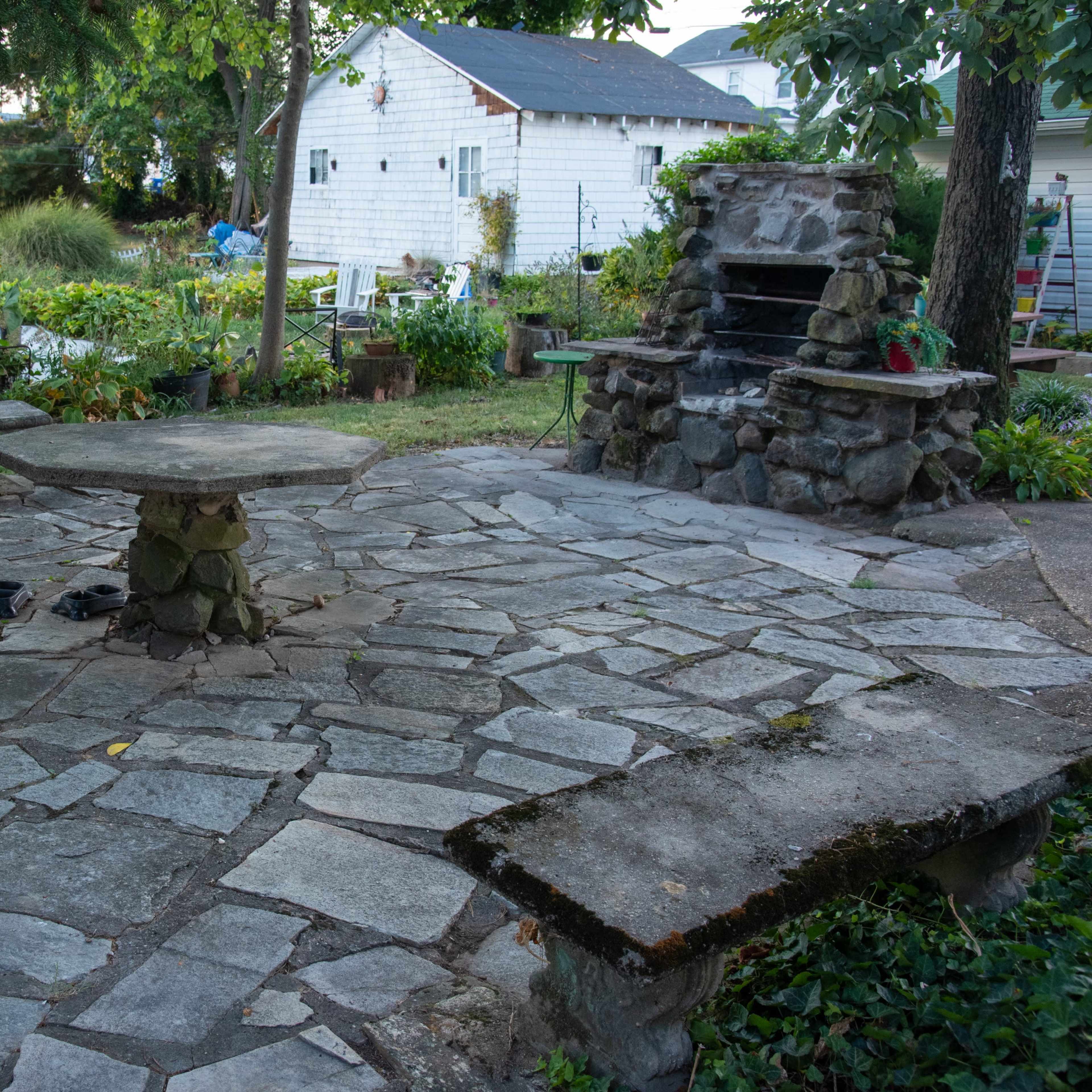 A stone patio features a central round table and a stone grill surrounded by benches in a garden setting.