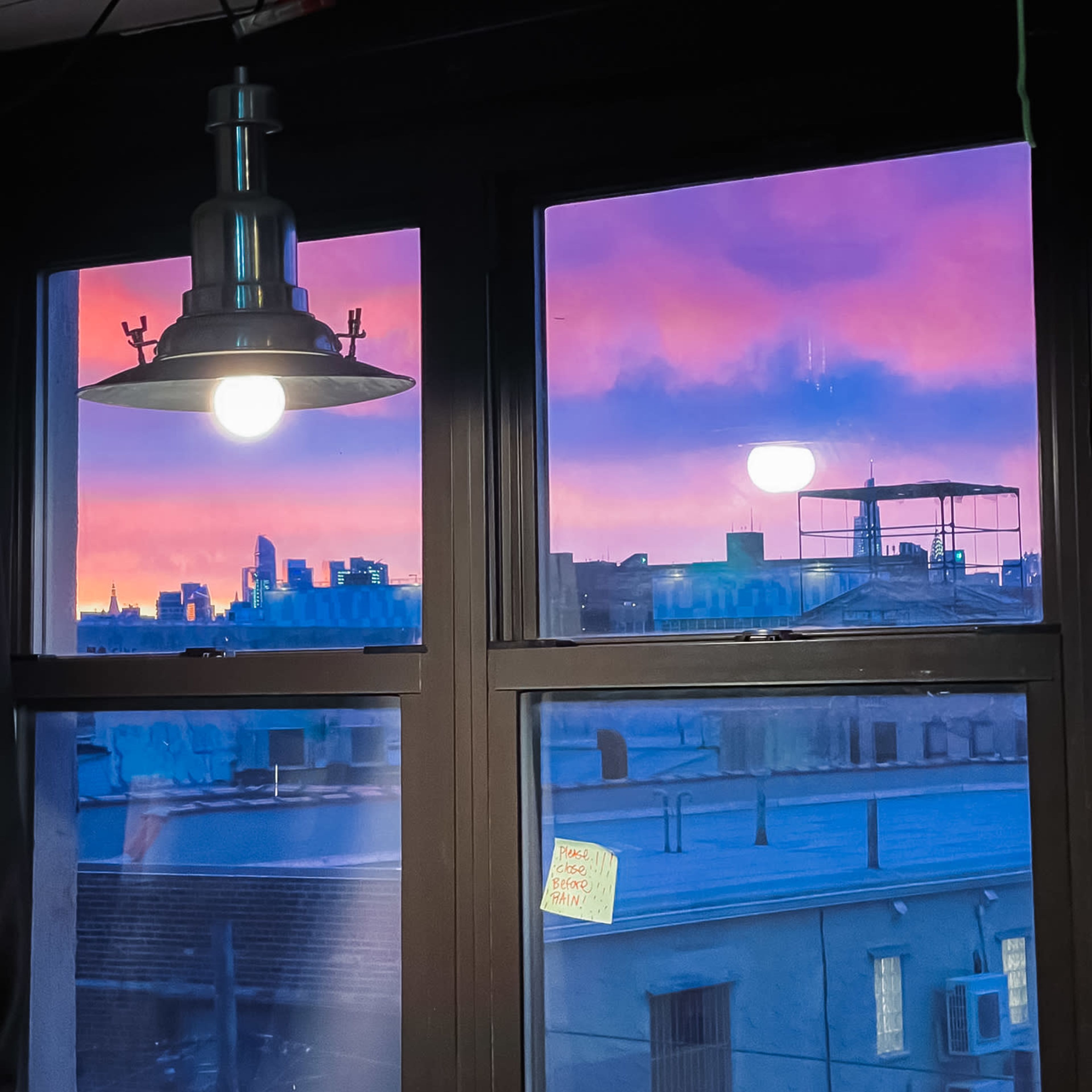 A pendant light hangs above a window, which frames a colorful sunset skyline and neighboring rooftops.