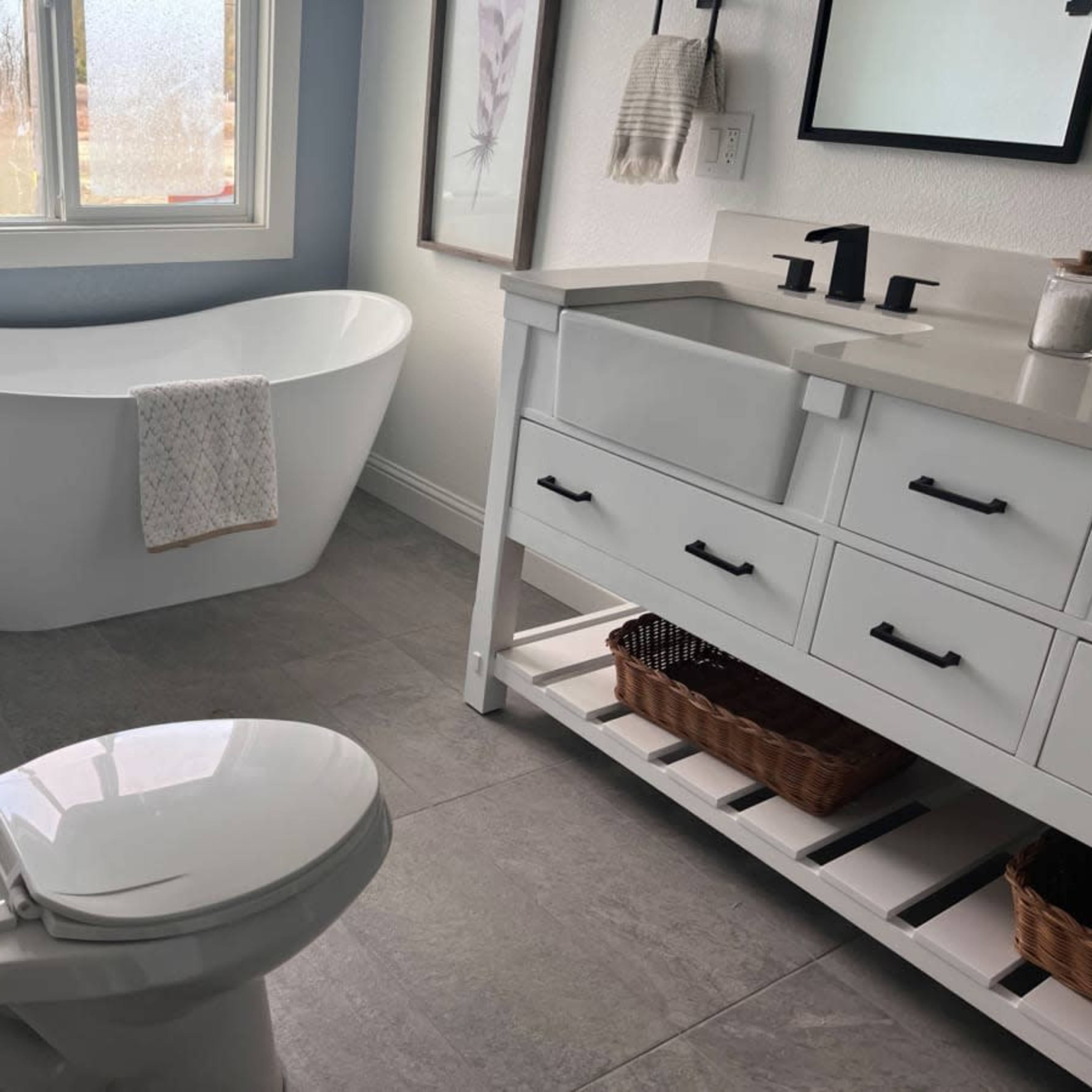 A modern bathroom with a freestanding bathtub, a vanity with drawers, and a toilet, all set against a light-colored wall and large window.