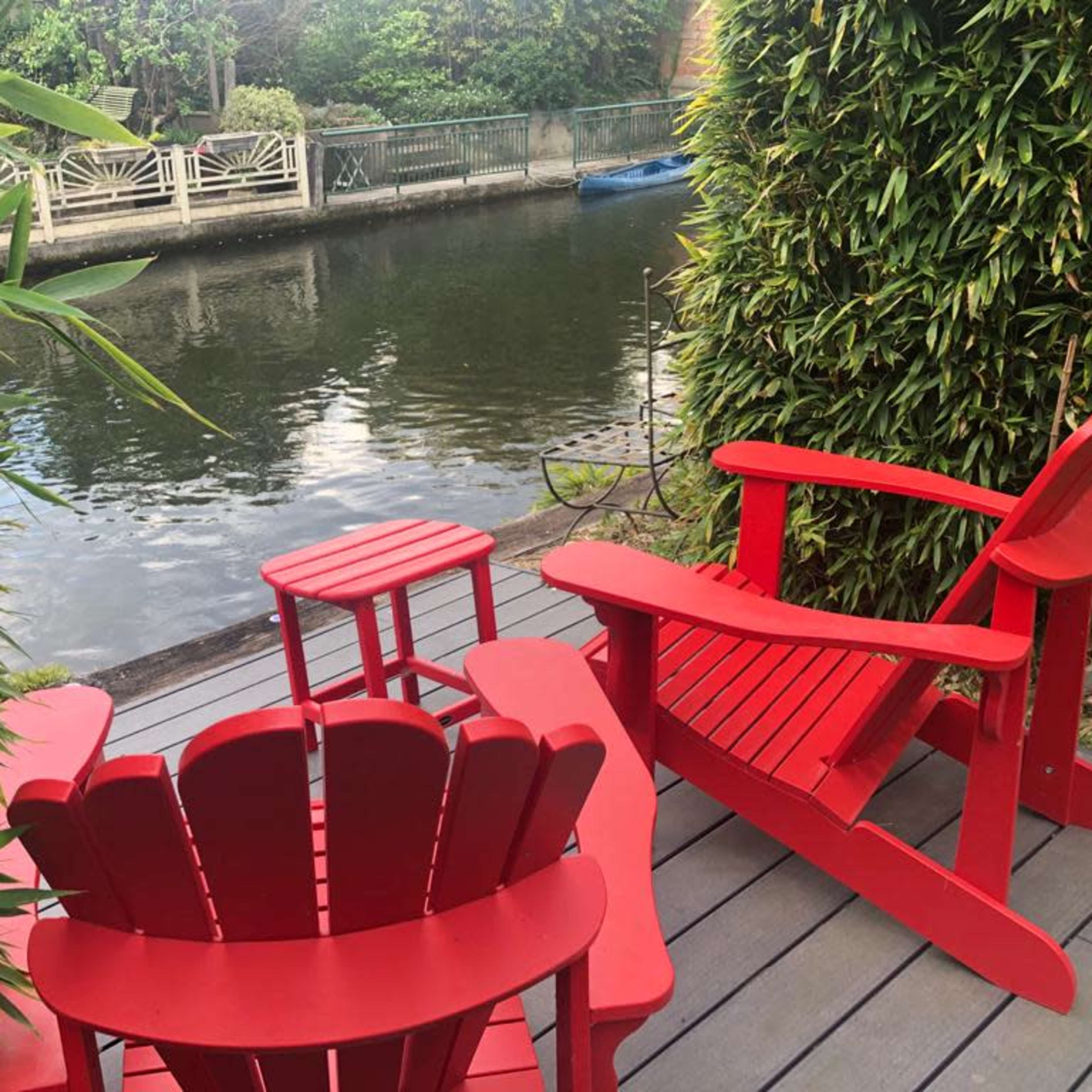 Red Adirondack chairs are arranged on a wooden deck overlooking a calm waterway surrounded by greenery.