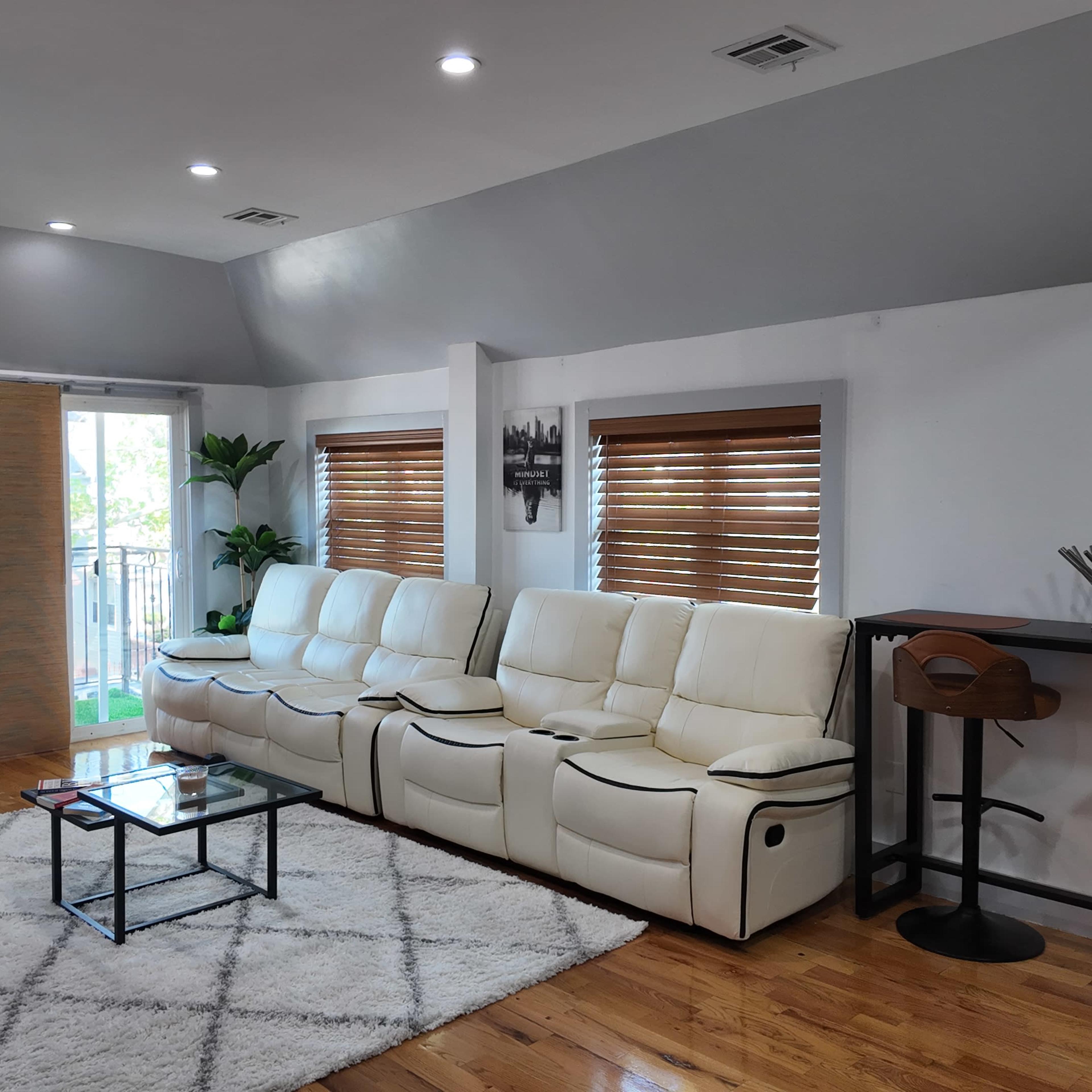 A modern living room features a white sectional sofa, a glass coffee table, and wooden flooring with large windows allowing natural light.