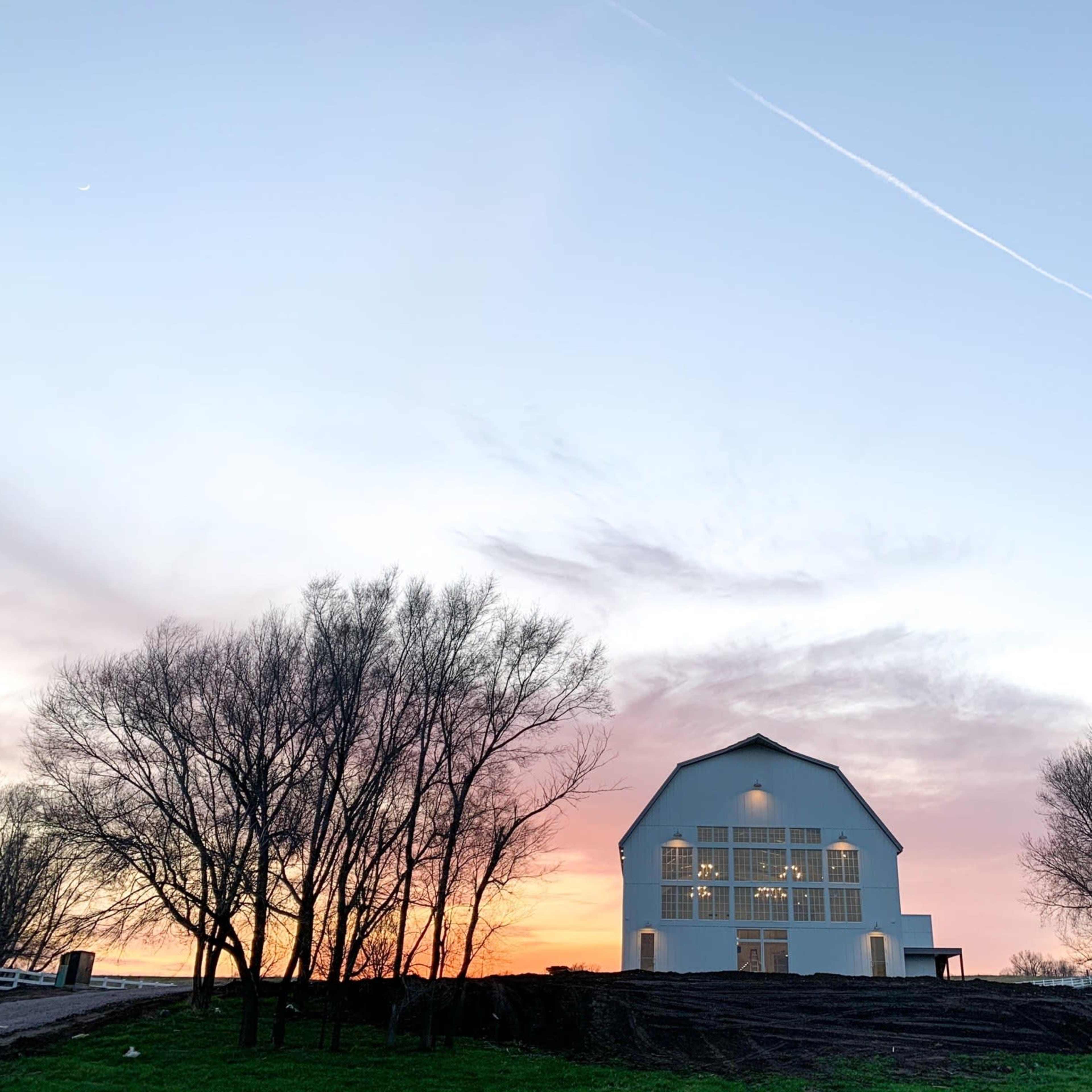 A white barn stands against a colorful sunset sky, framed by bare trees.