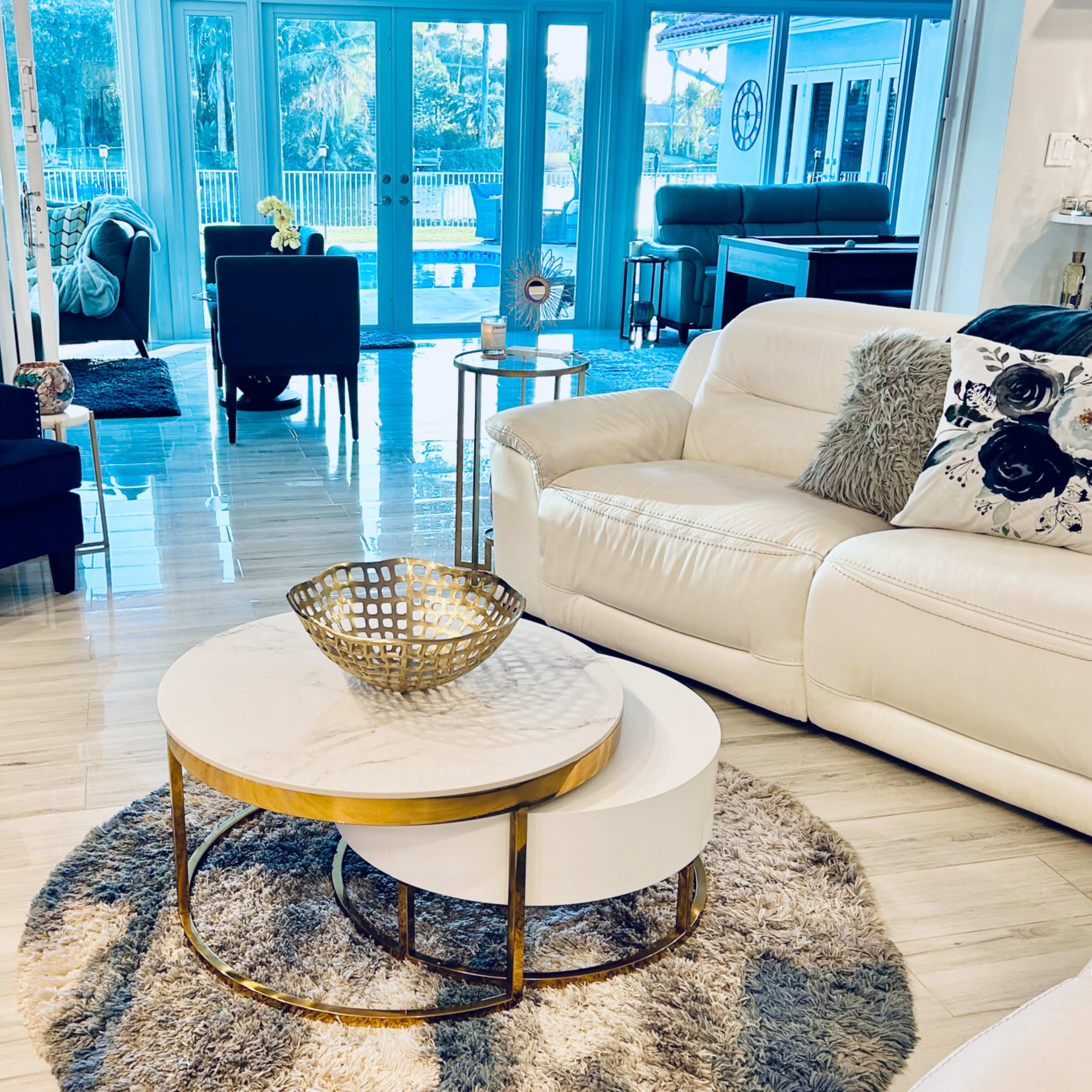 The living room features a white leather sofa, a round coffee table, a plush rug, and large windows overlooking a patio with outdoor furniture.