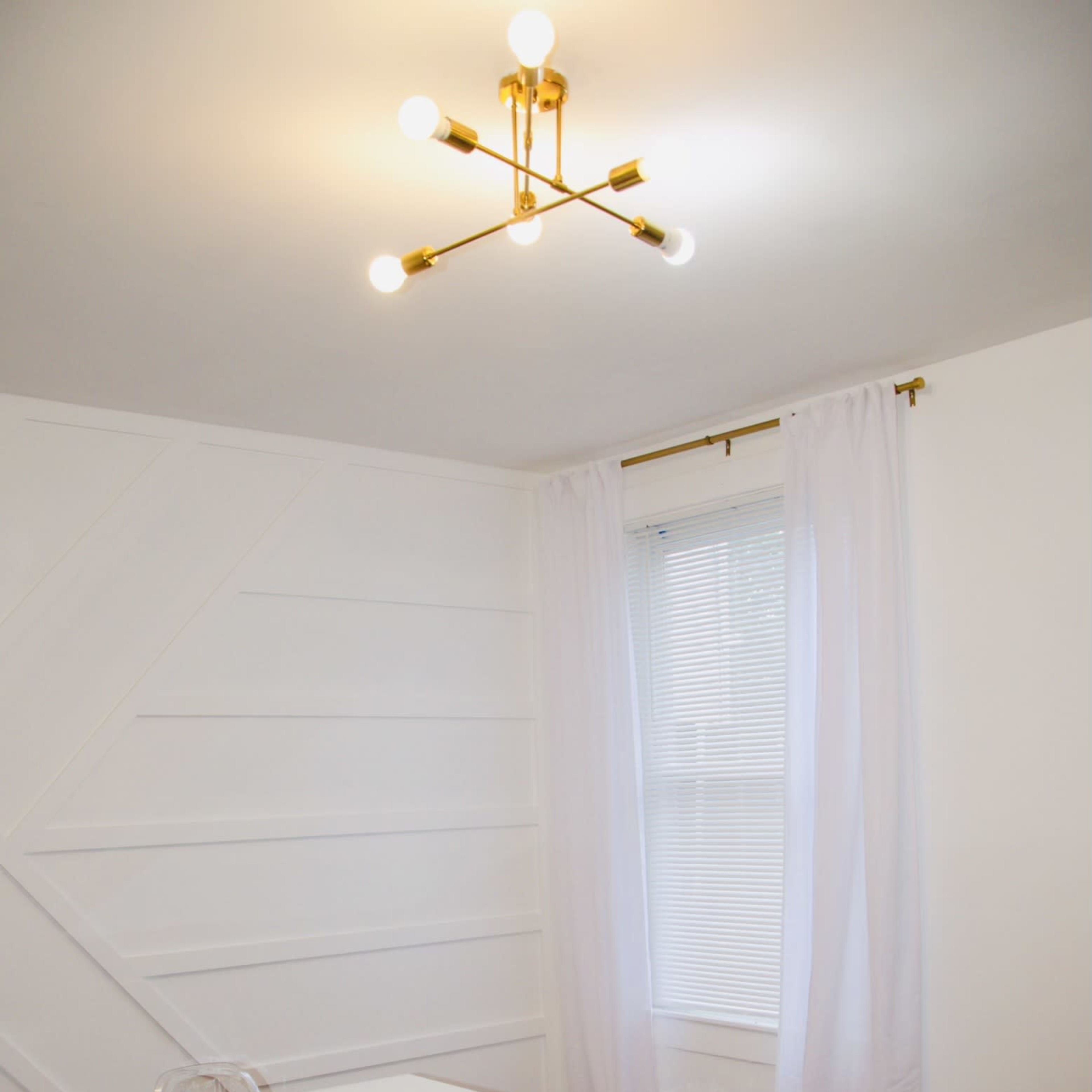 A modern dining room features a white table surrounded by clear chairs under a geometric ceiling light and a window with sheer curtains.