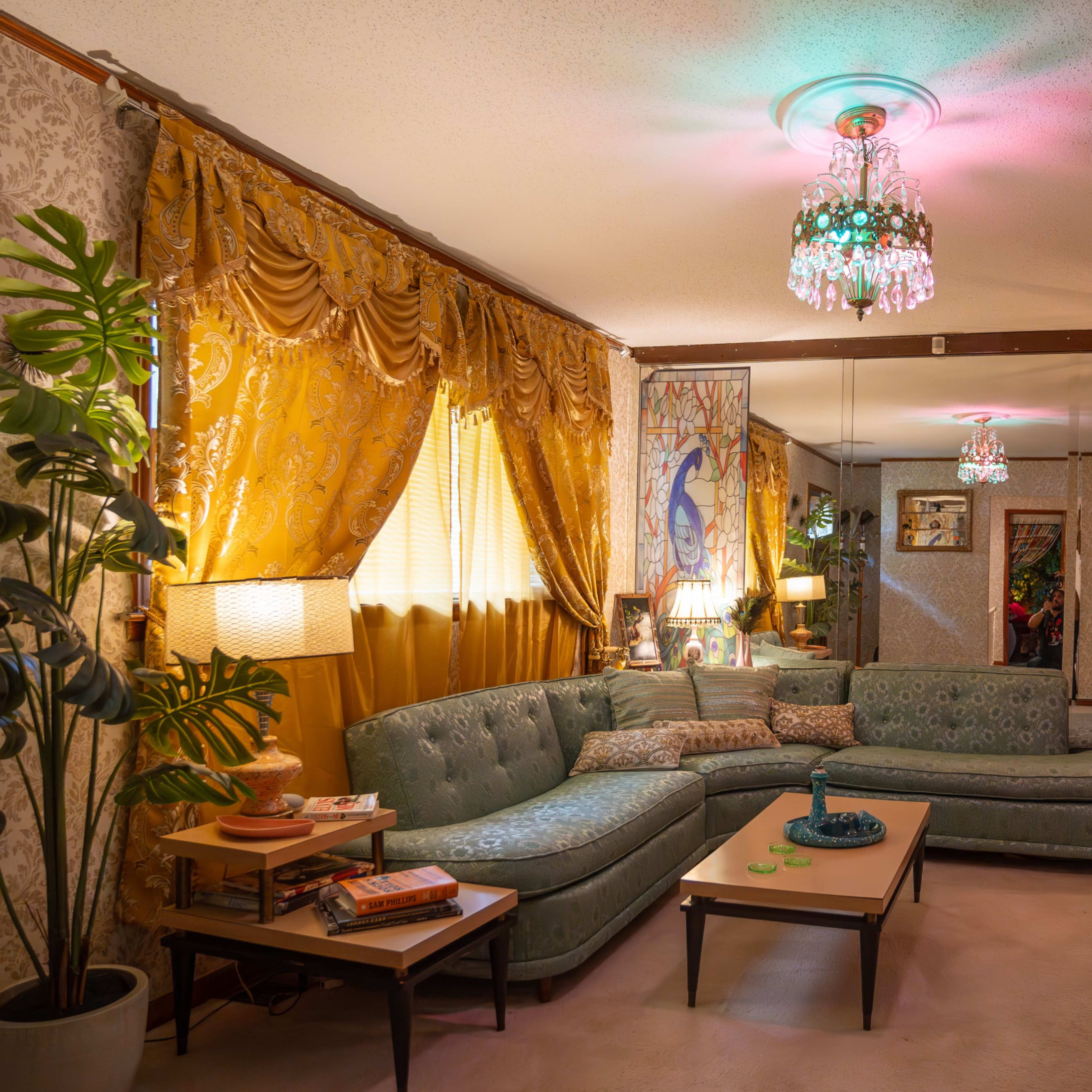 A retro living room features a turquoise sectional couch, patterned walls, and colorful chandeliers providing ambient light.
