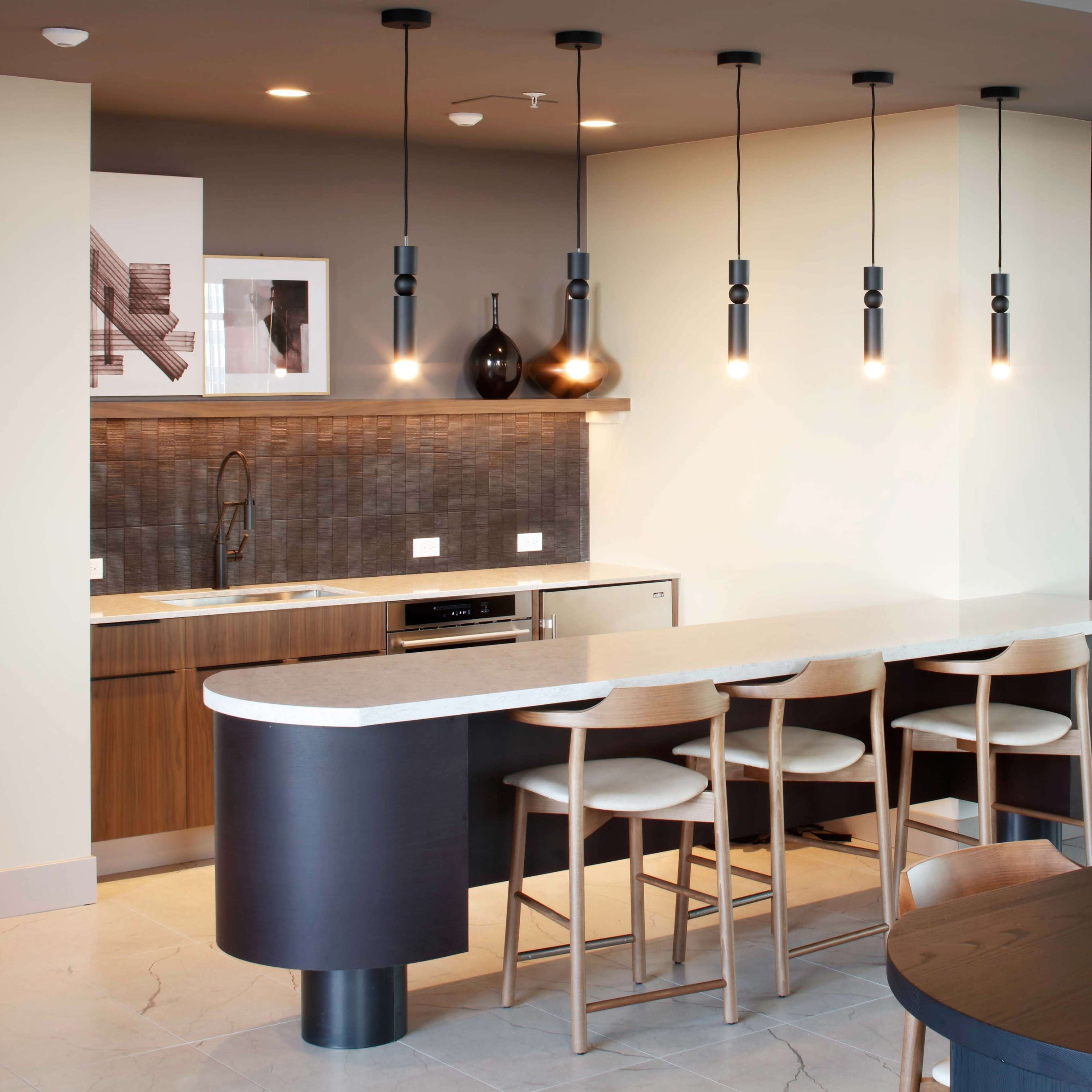 A modern kitchen area with a bar counter, four stools, pendant lights, and an adjacent dining table, featuring wooden cabinetry and a sleek countertop.