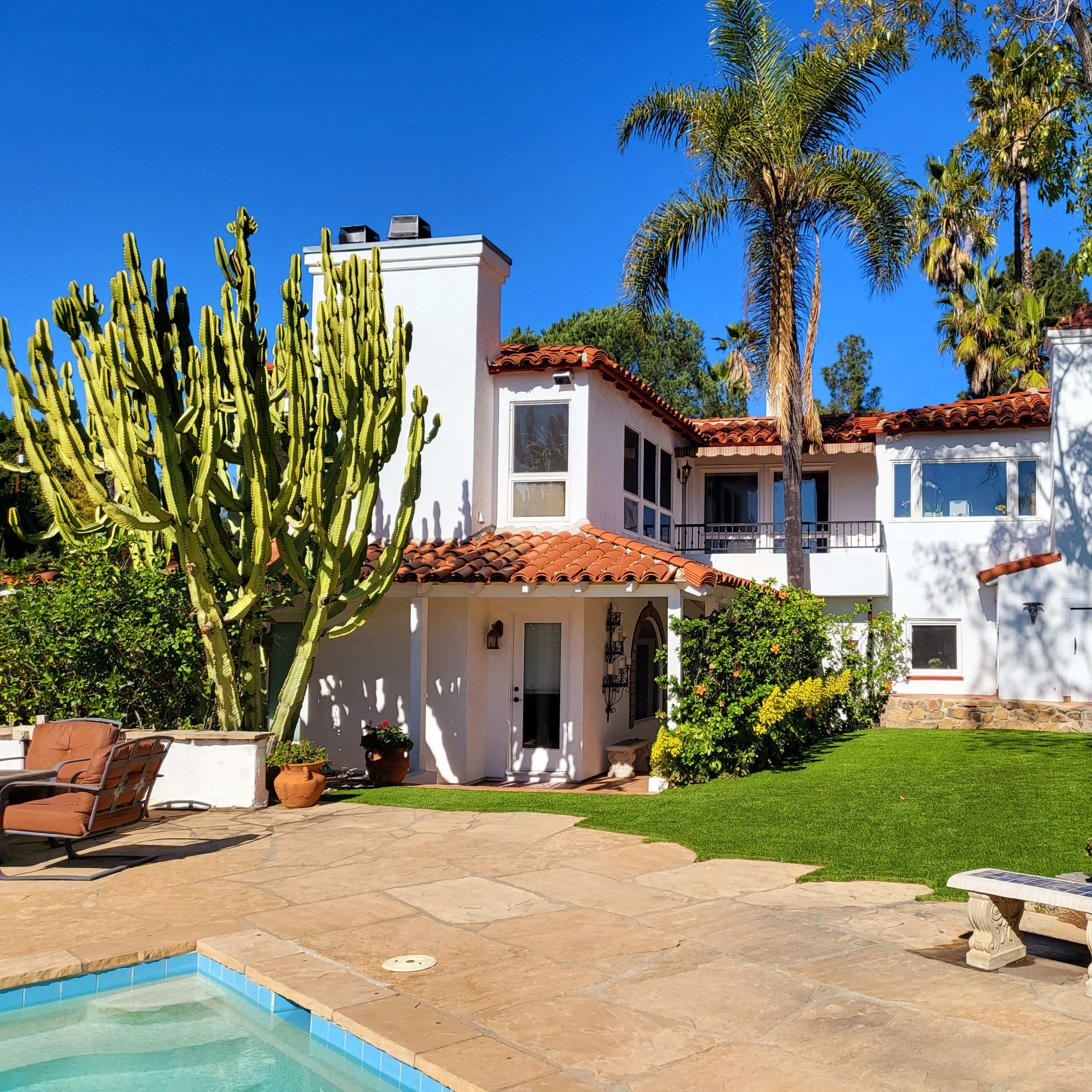 A two-story house with a red tile roof, surrounded by lush greenery and a swimming pool in the foreground.
