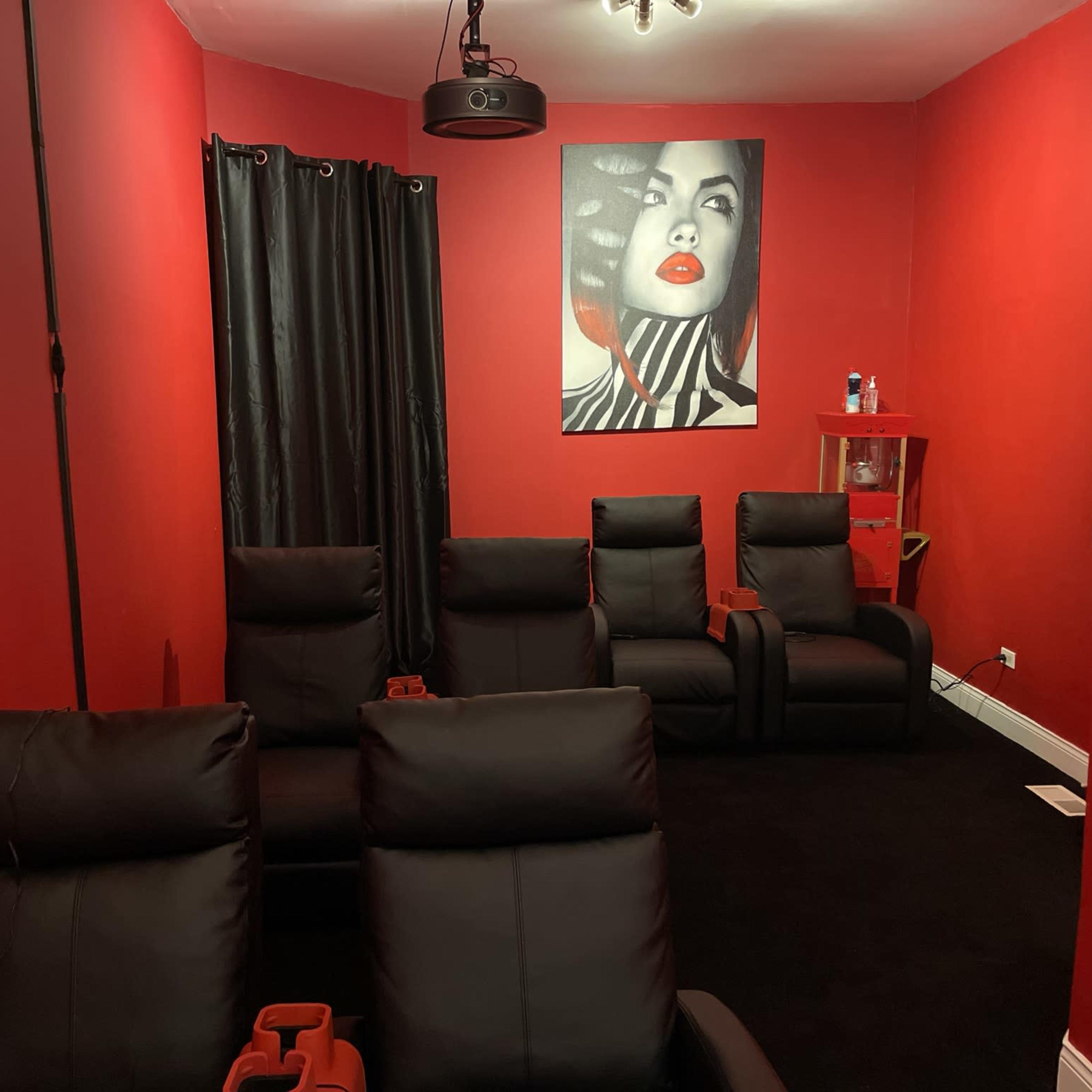 The room features black reclining chairs facing a wall adorned with a striking artwork of a woman, all set against red walls and black curtains.