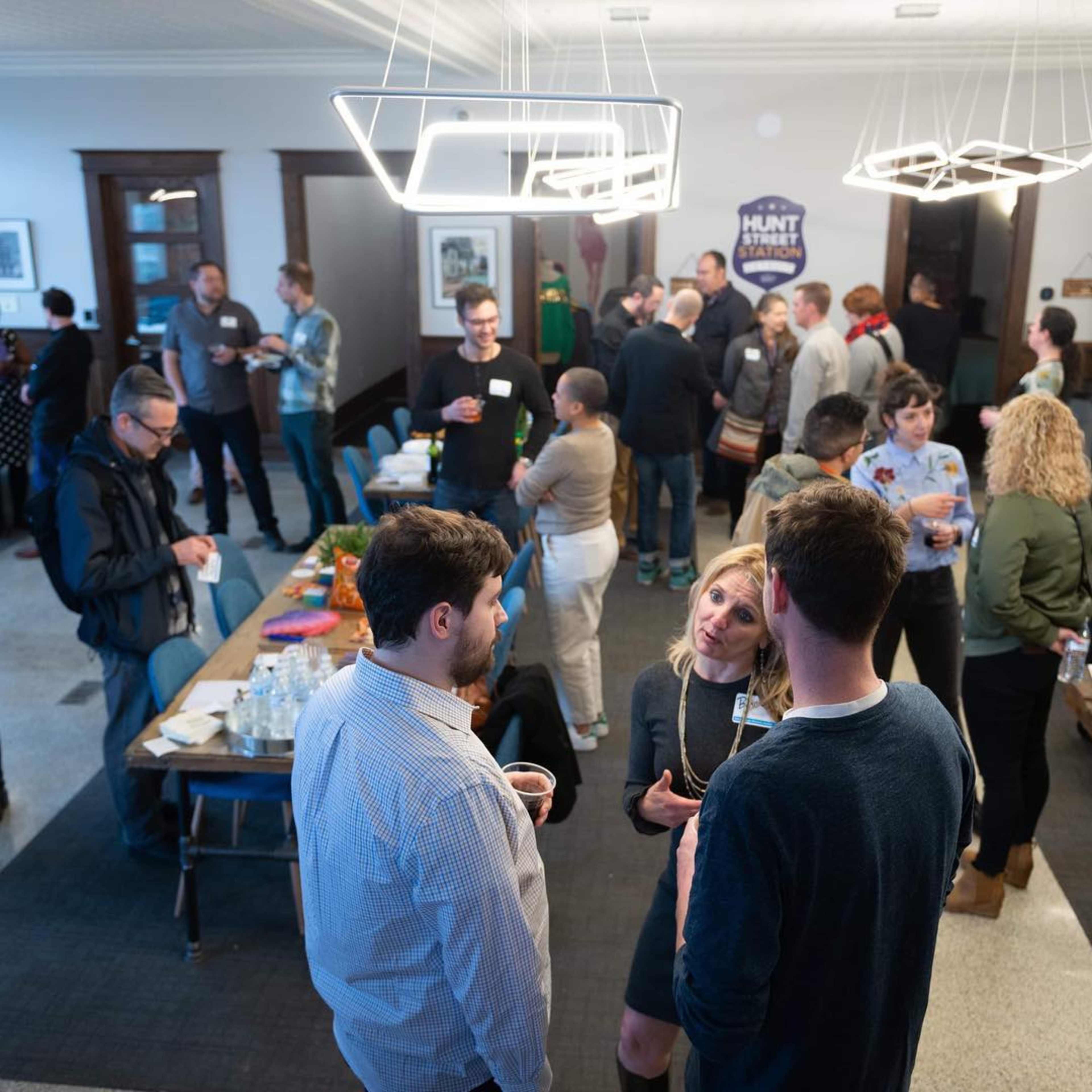 A networking event is taking place in a room with people mingling, seated at tables, and conversation happening among attendees.