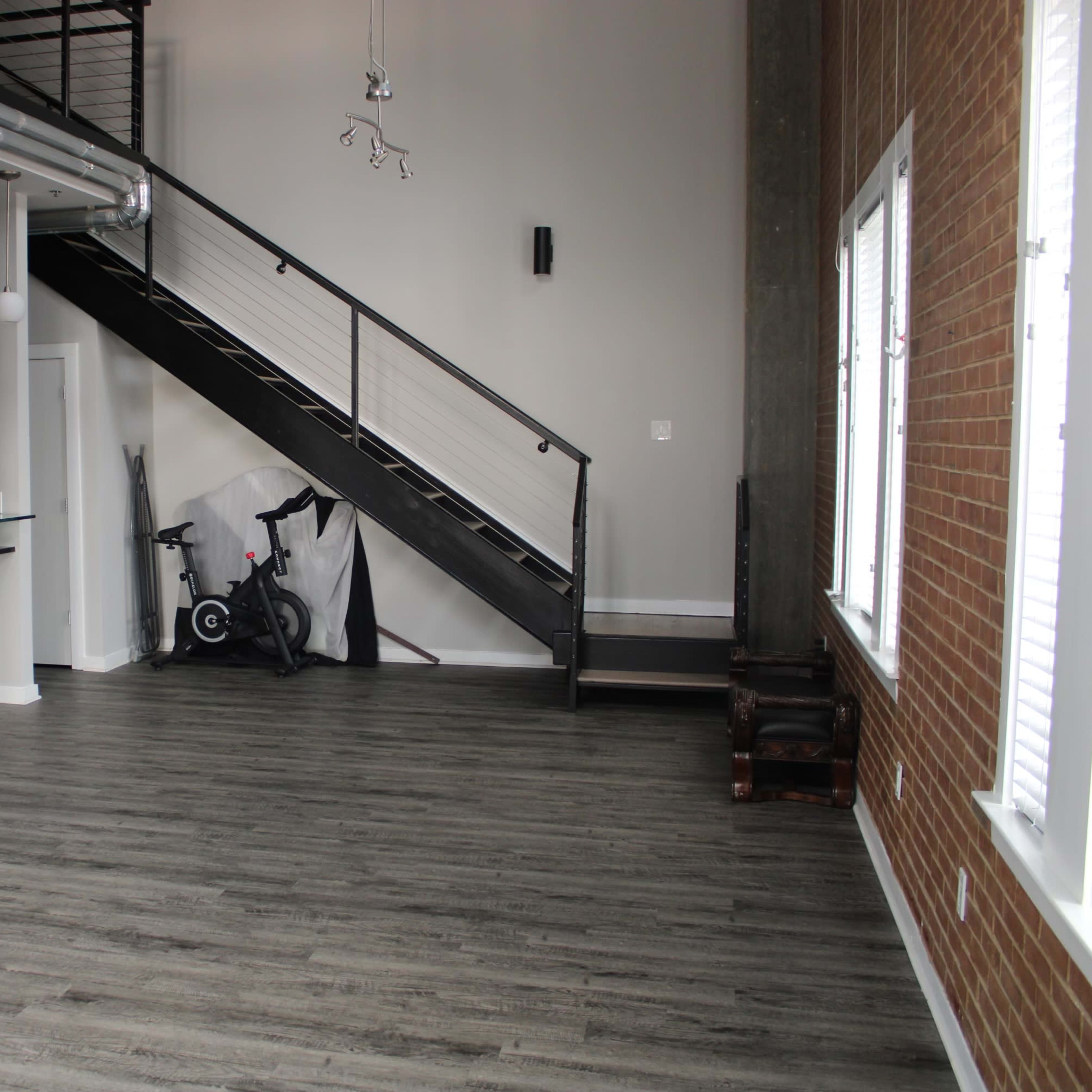 An empty industrial-style living space featuring a staircase, large windows, and a corner with exercise equipment.