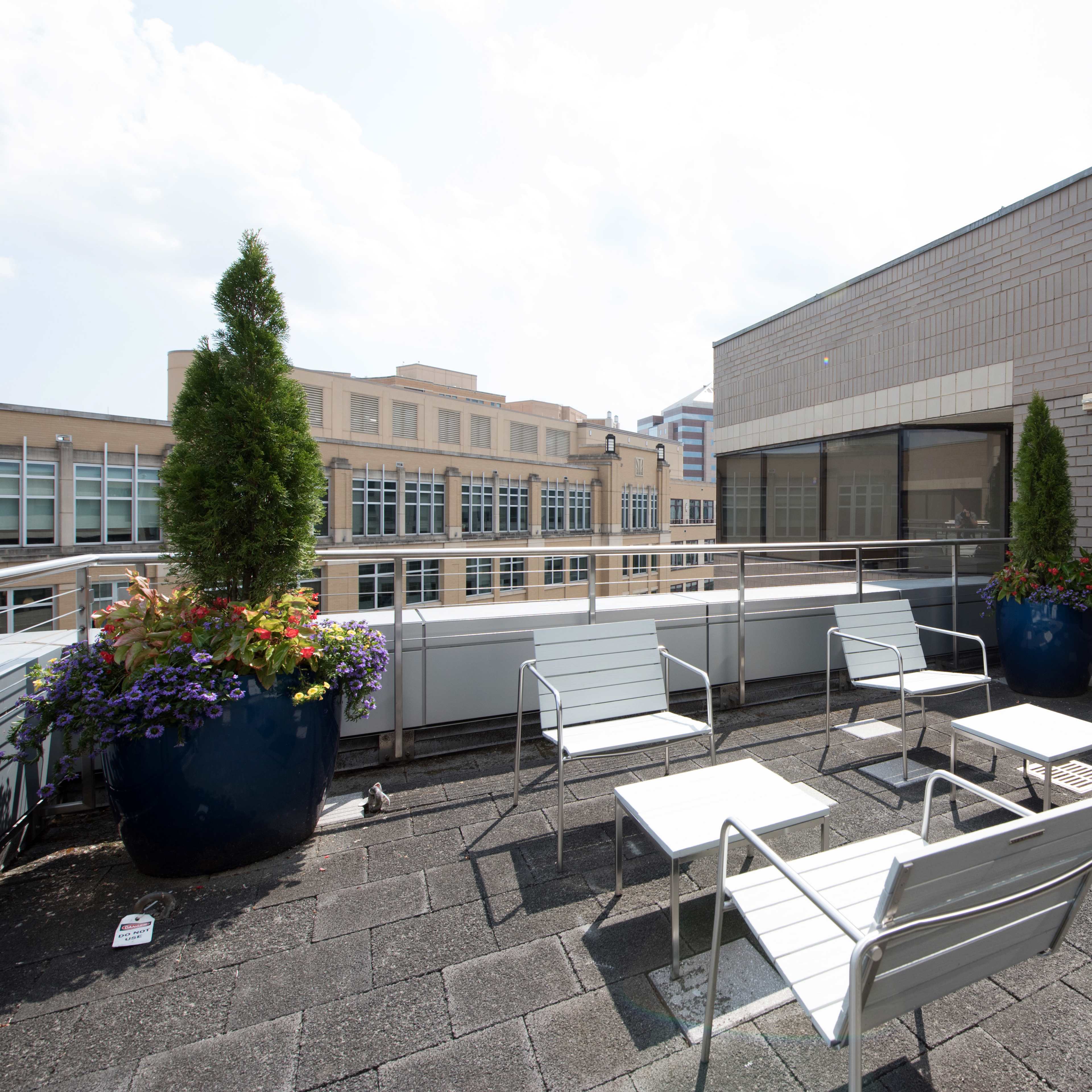 A rooftop terrace features metal chairs and tables surrounded by large planters filled with flowers.