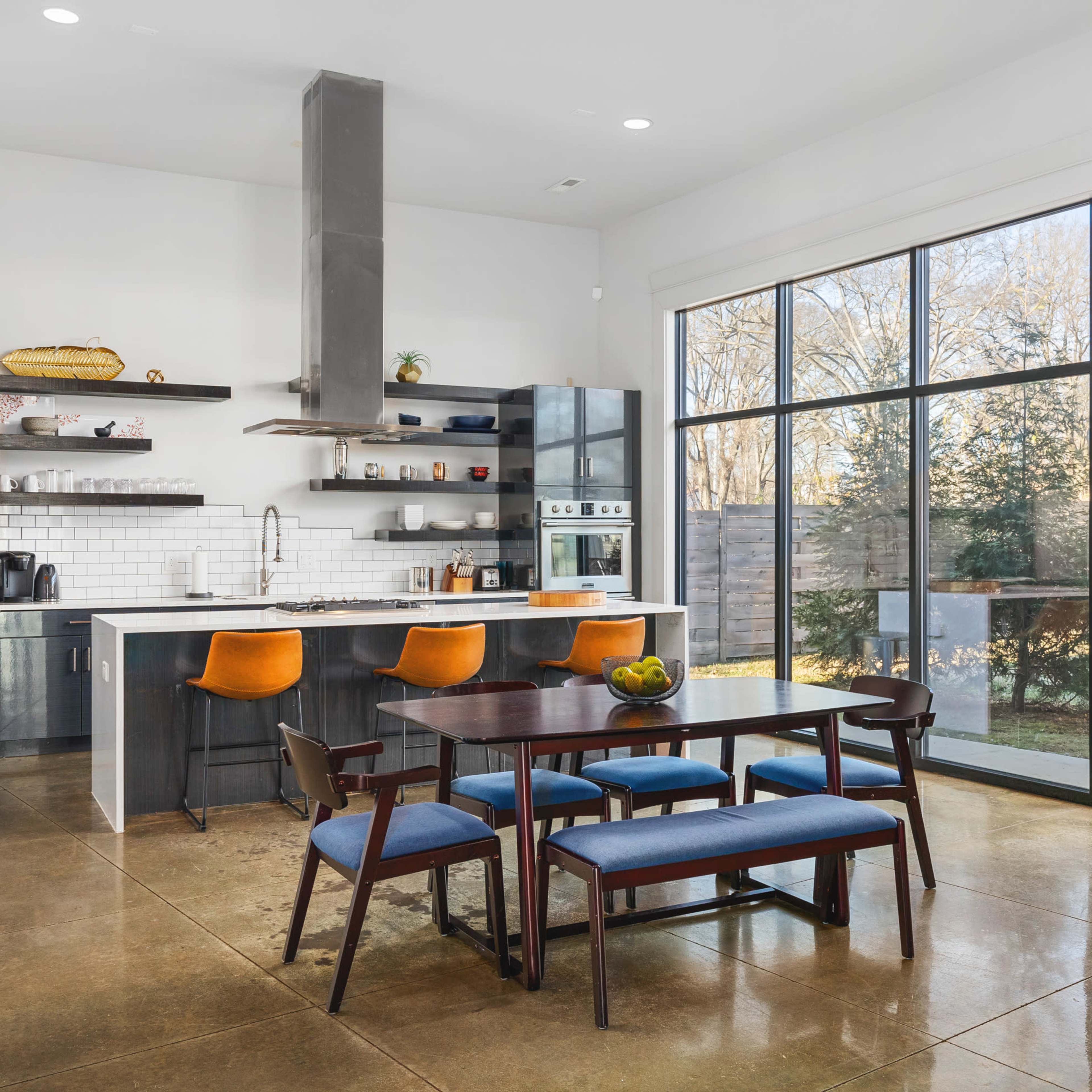 A modern kitchen features a large island with bar stools, a dining table, and a wall of windows providing natural light.