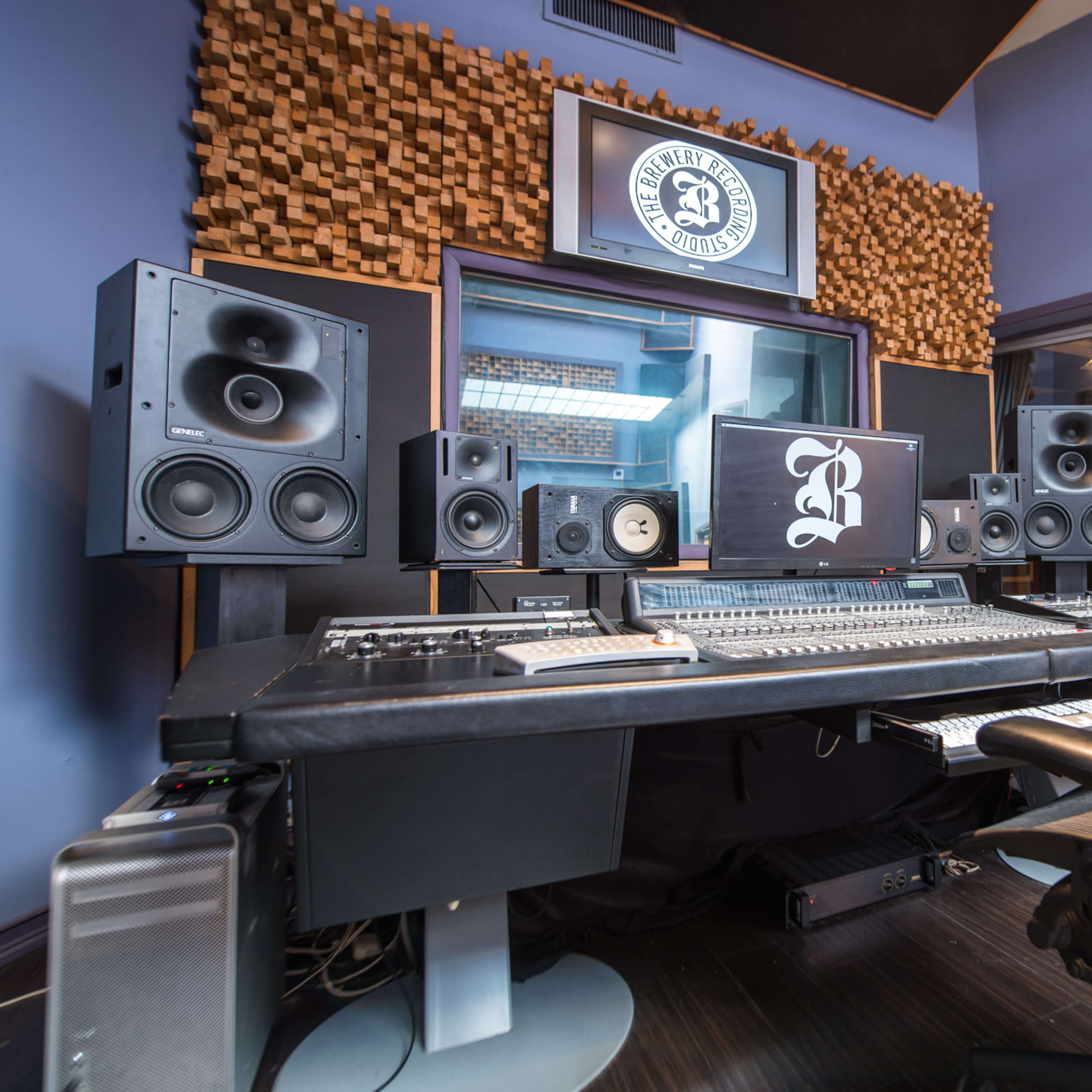 A music recording studio features a large console surrounded by multiple speakers and a computer monitor displaying a logo on the wall behind.