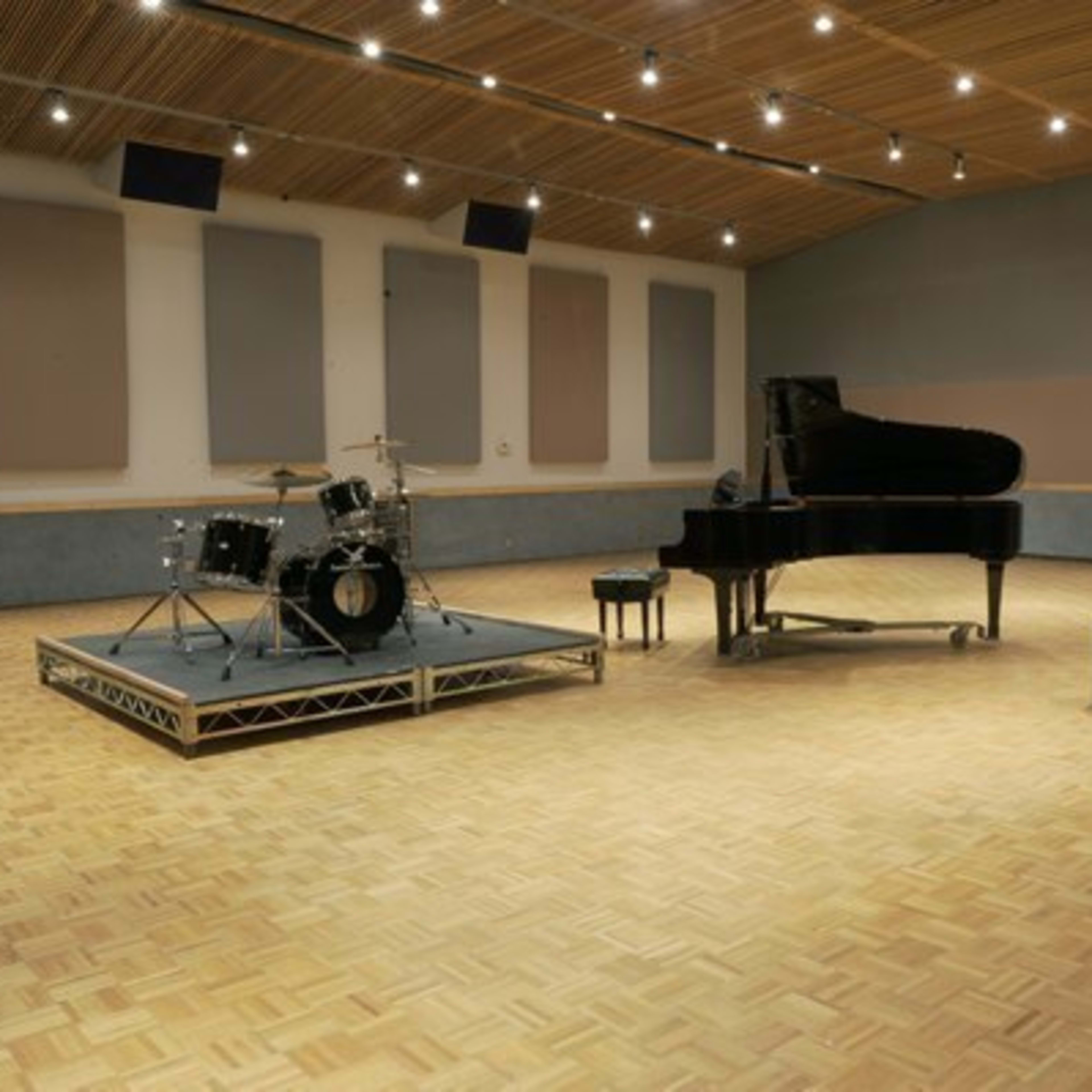 A spacious recording studio featuring a grand piano, a drum set on a raised platform, an electric guitar, and an amplifier.