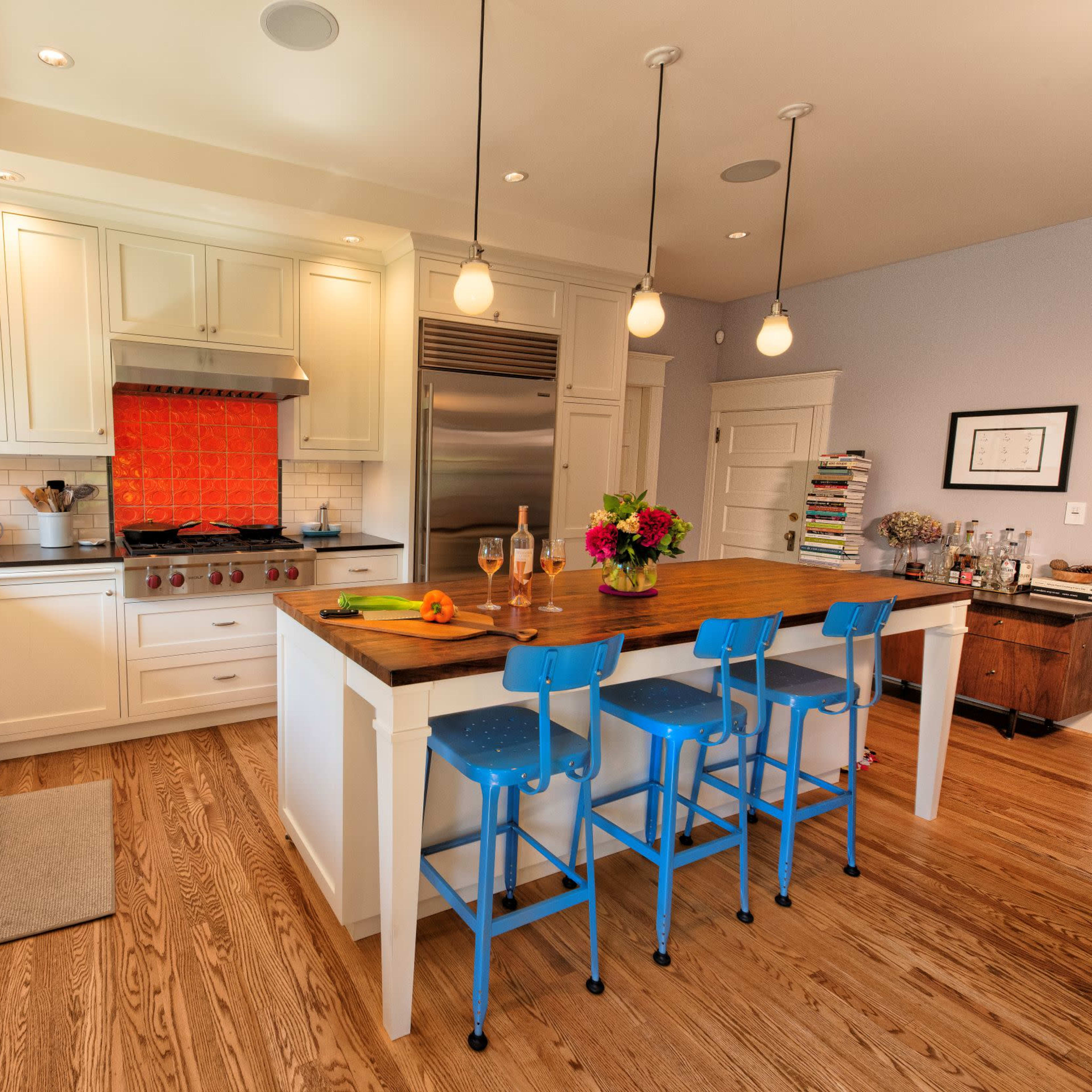 A modern kitchen features white cabinets, a large island with blue stools, and an orange tile backsplash behind the stove.