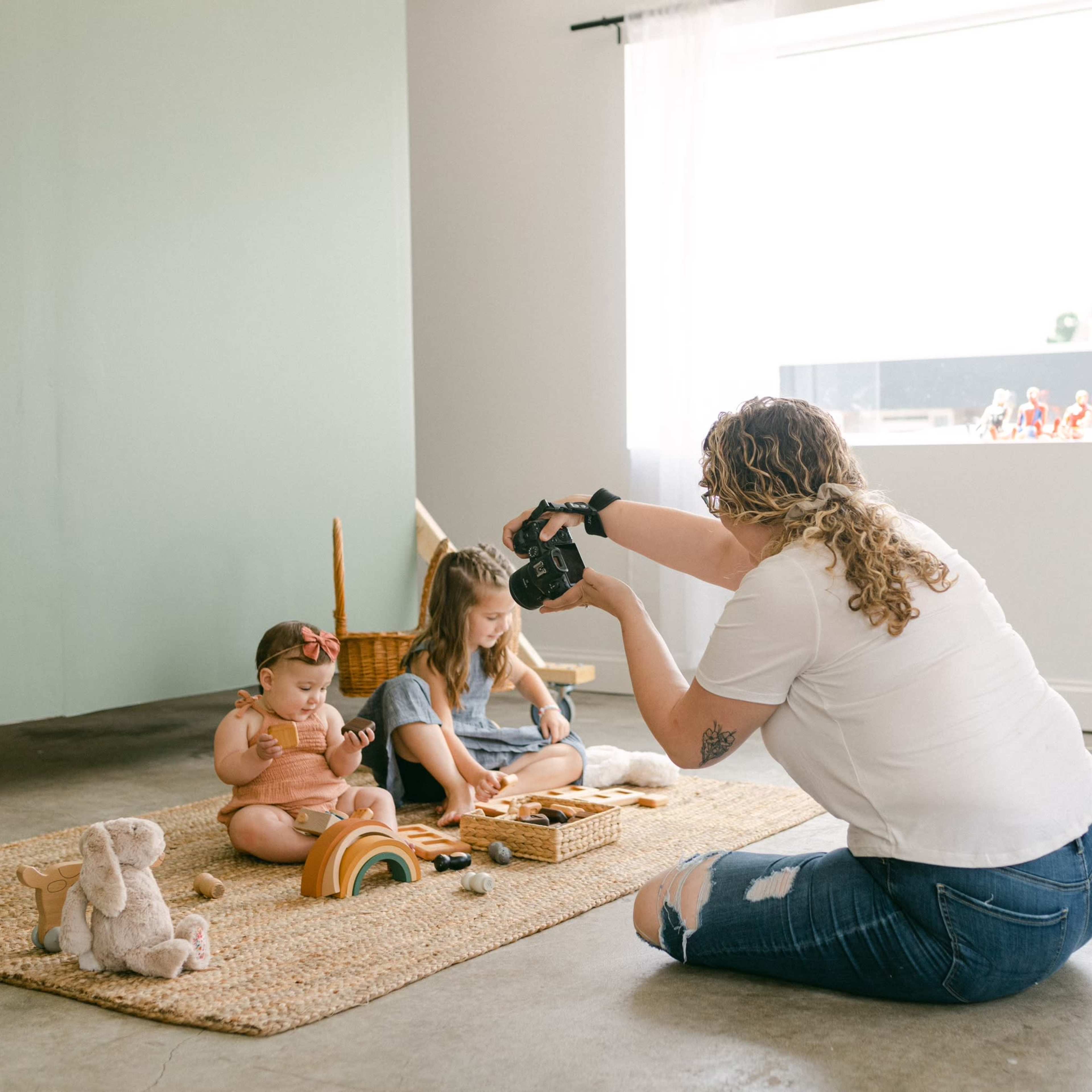 A woman photographs two children playing on a rug in a brightly lit room.