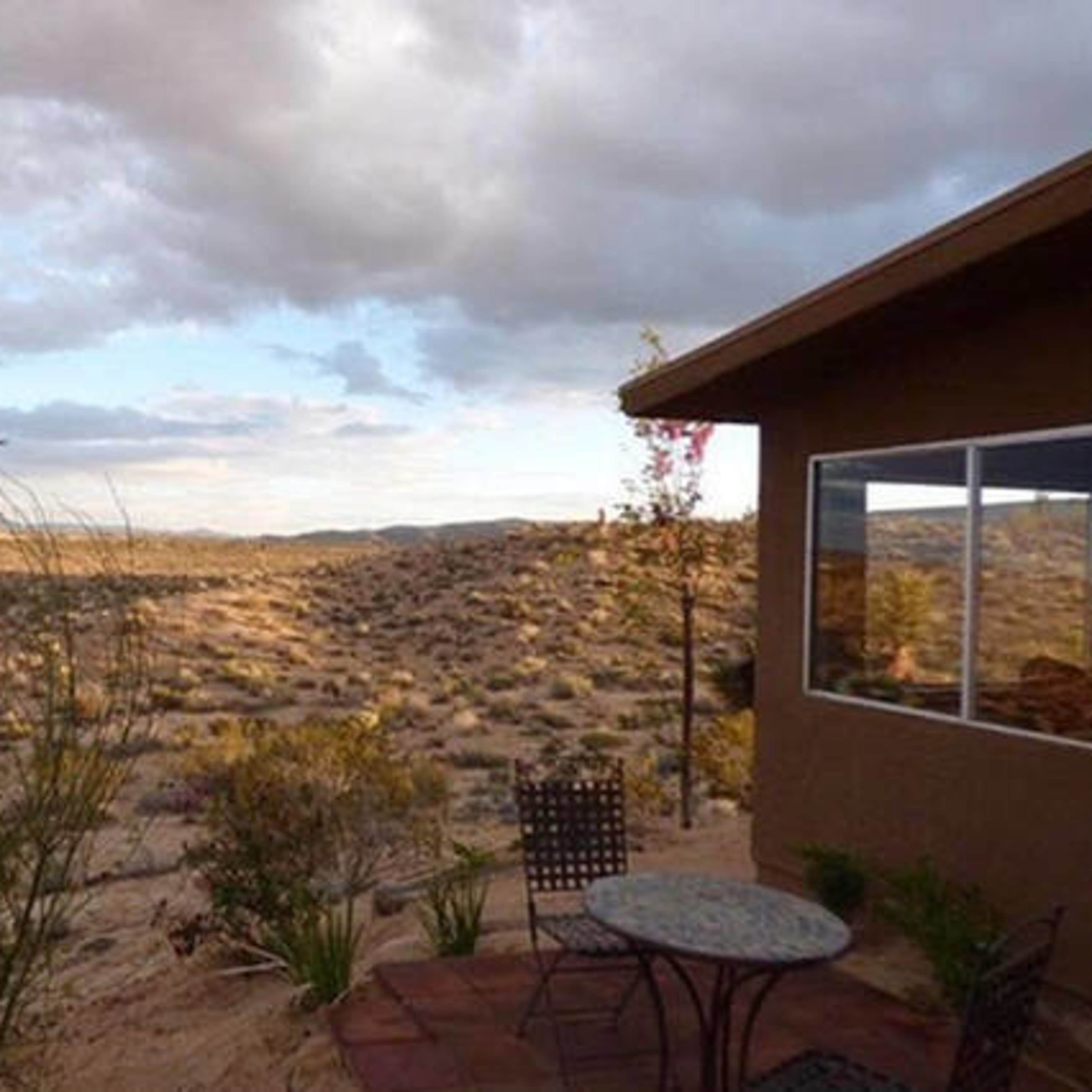 A desert landscape with sparse vegetation and a house in the foreground, featuring a small outdoor table and chairs.