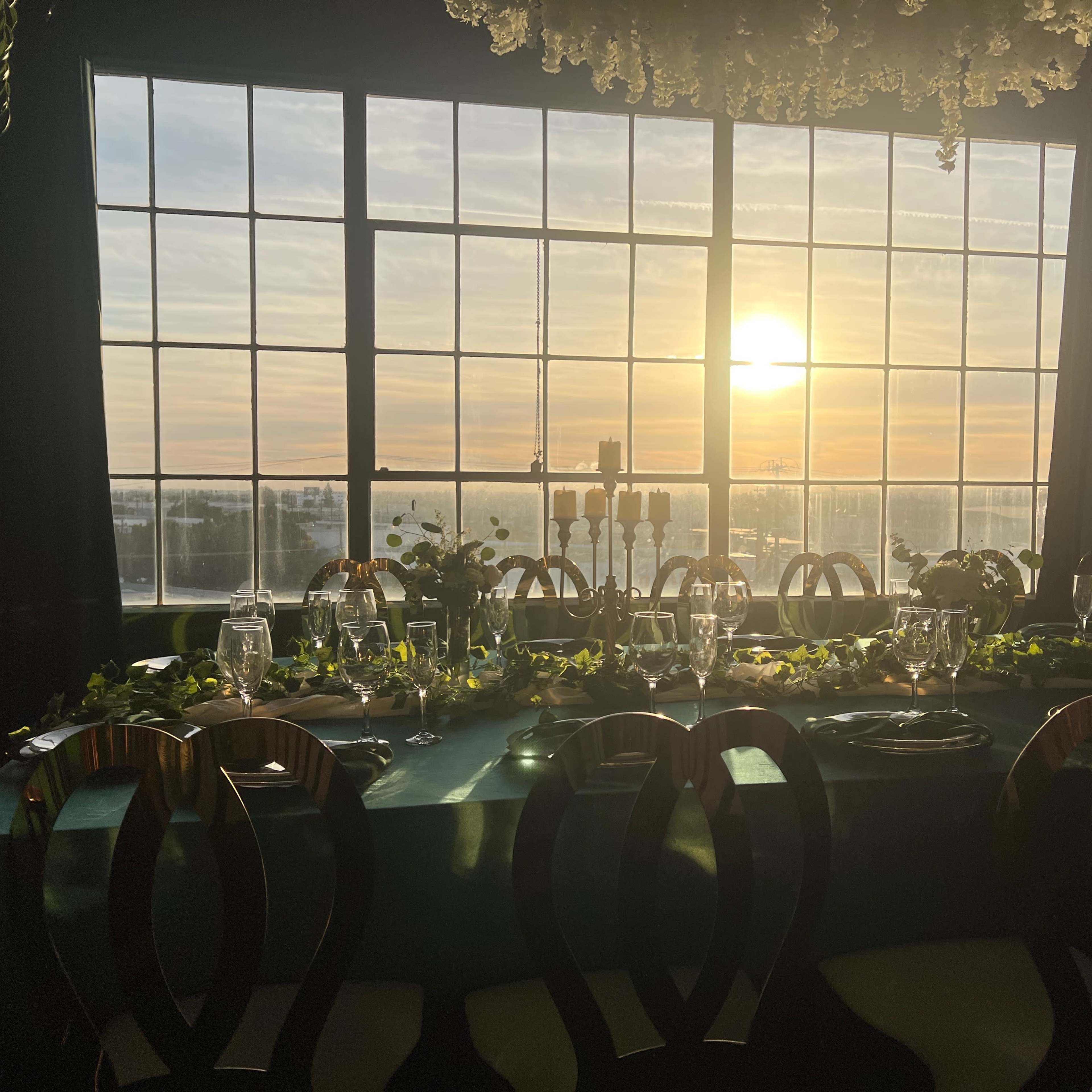 A beautifully arranged dining table featuring elegant table settings, with a backdrop of large windows displaying a sunset over a scenic landscape.