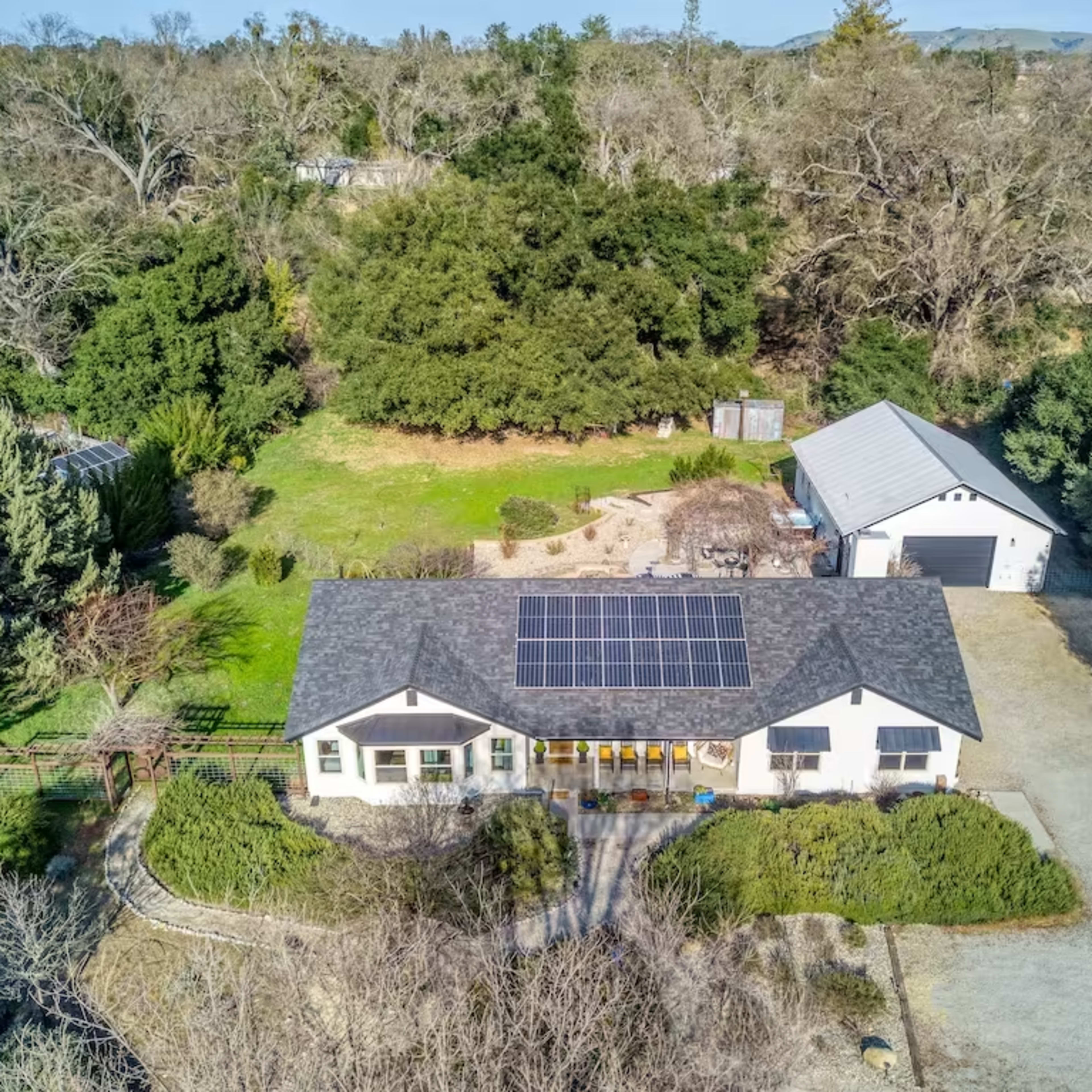 A single-story house with a solar panel roof is situated on a spacious lot surrounded by trees and a detached garage.