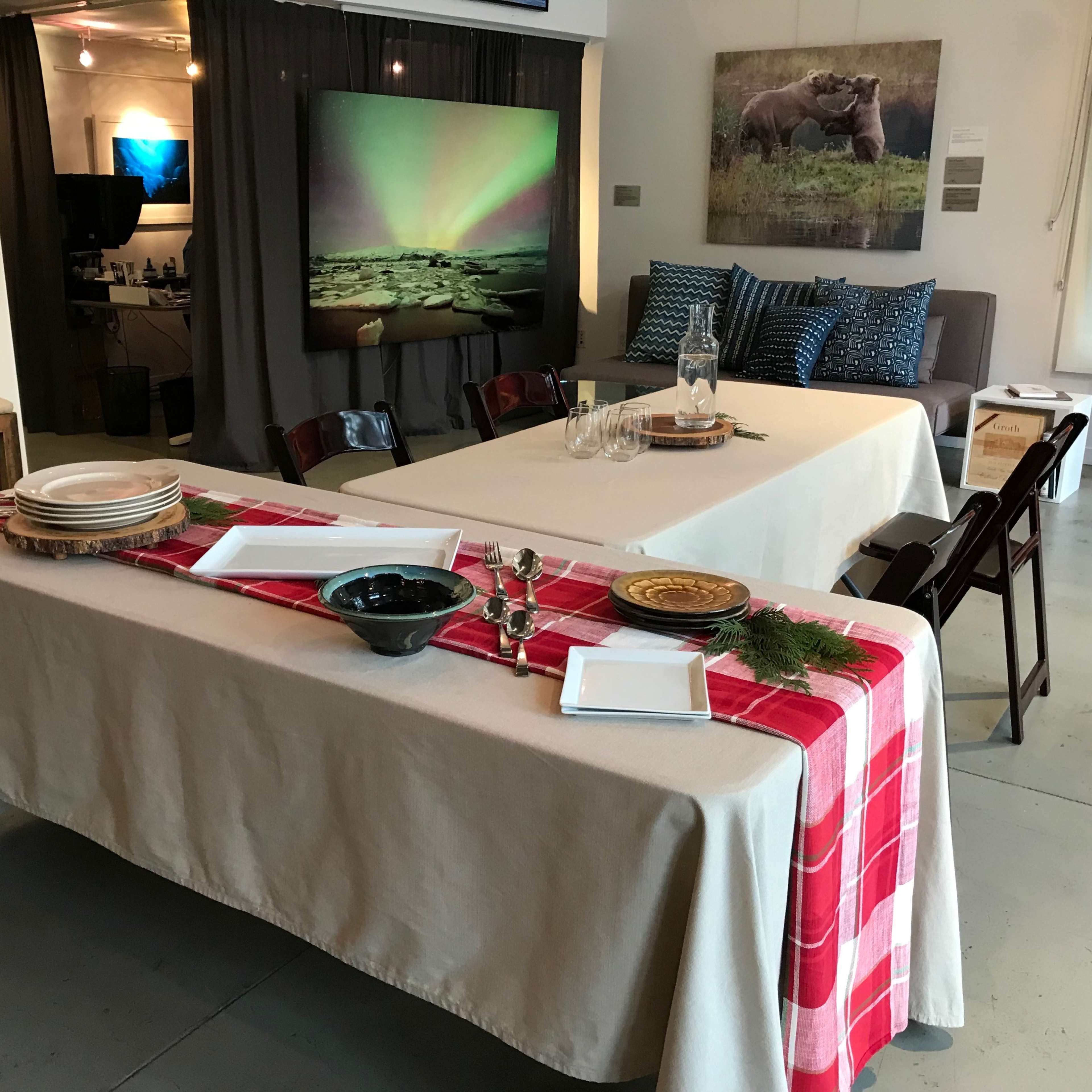 A dining area is set up with a long table covered in a beige cloth, featuring a red checkered runner, various dishes, and glassware, surrounded by art displayed on the walls.