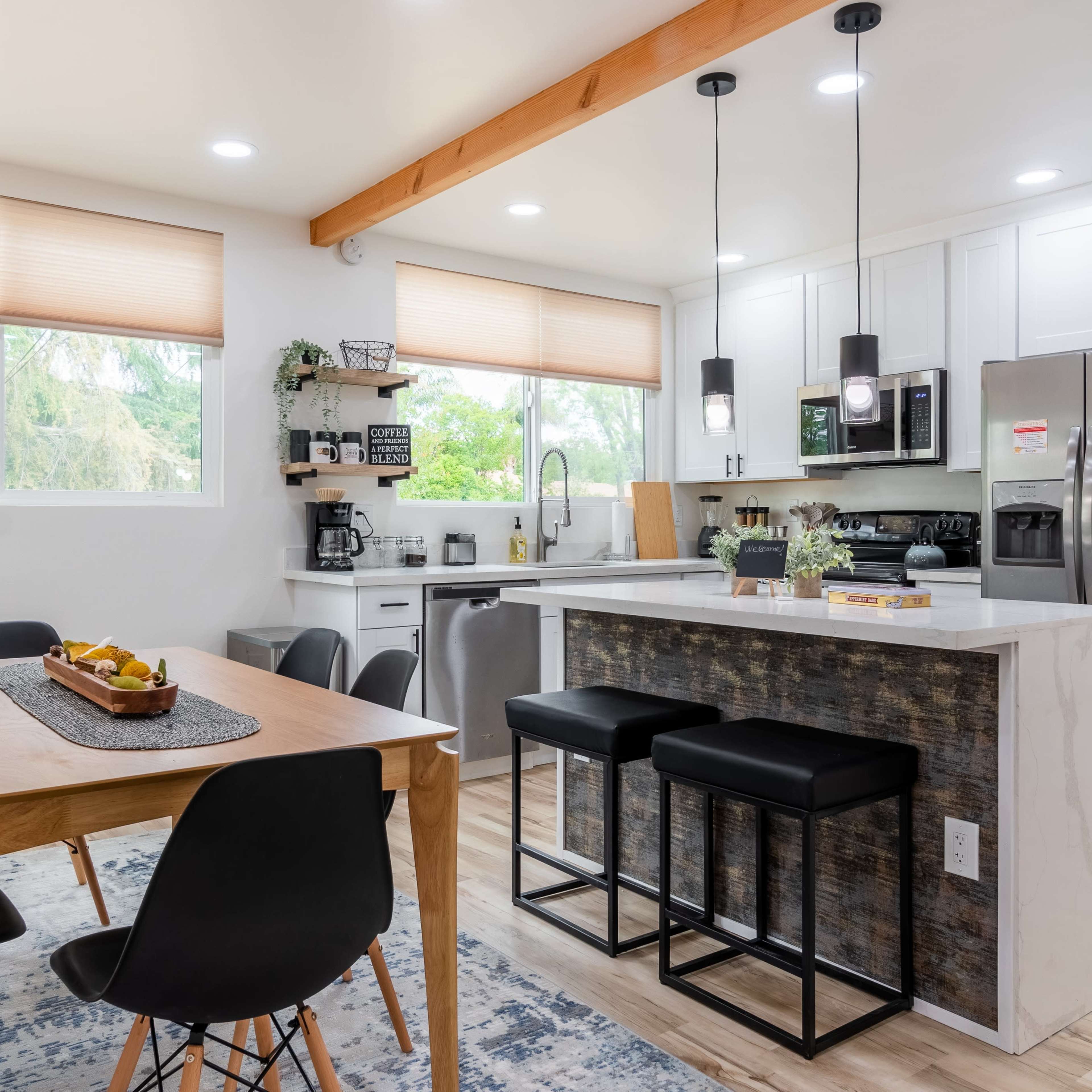 A modern kitchen and dining area featuring a wooden table with black chairs, a countertop with bar stools, and large windows allowing natural light to enter.