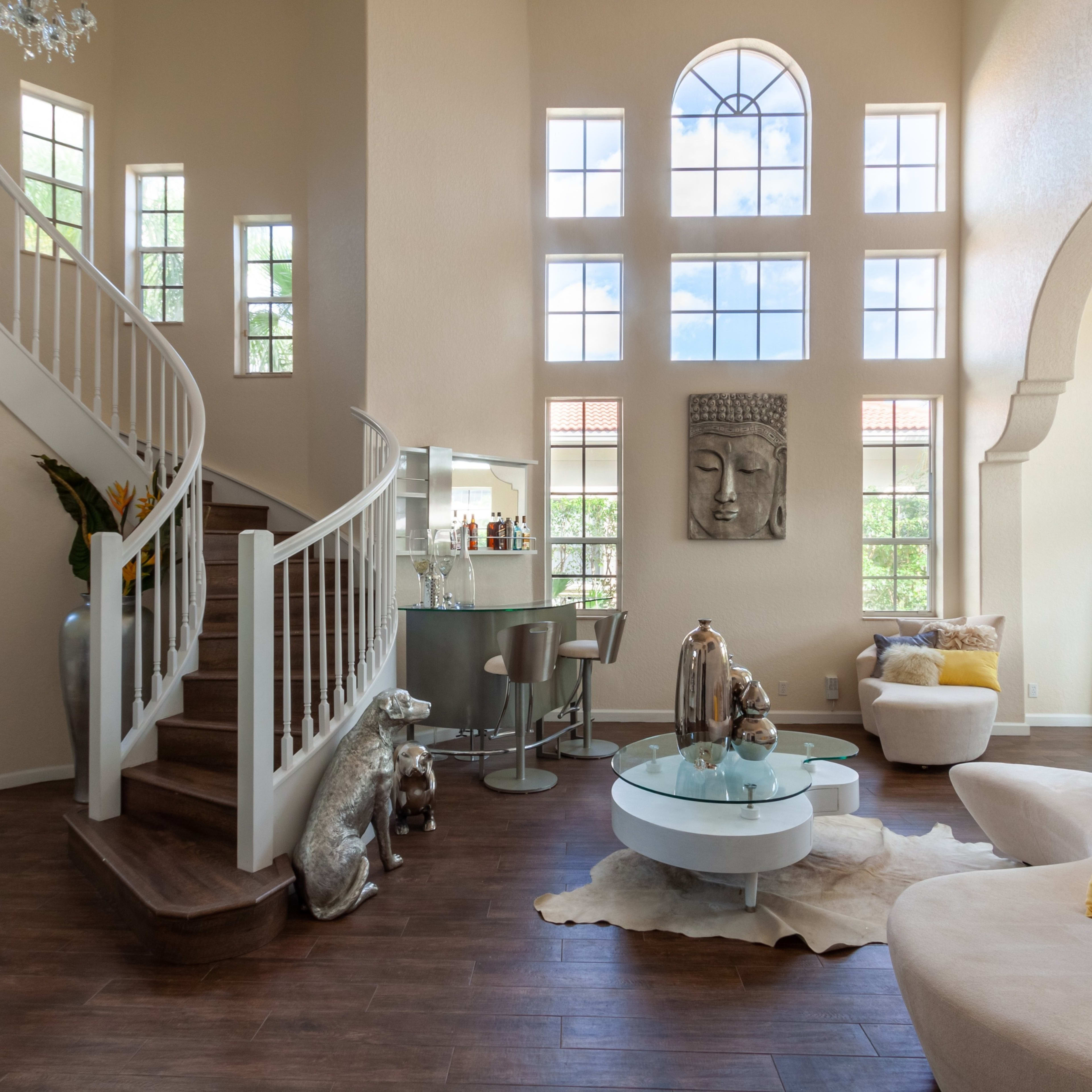 A spacious living room features large windows, a curved staircase, and modern furniture arranged around a coffee table.
