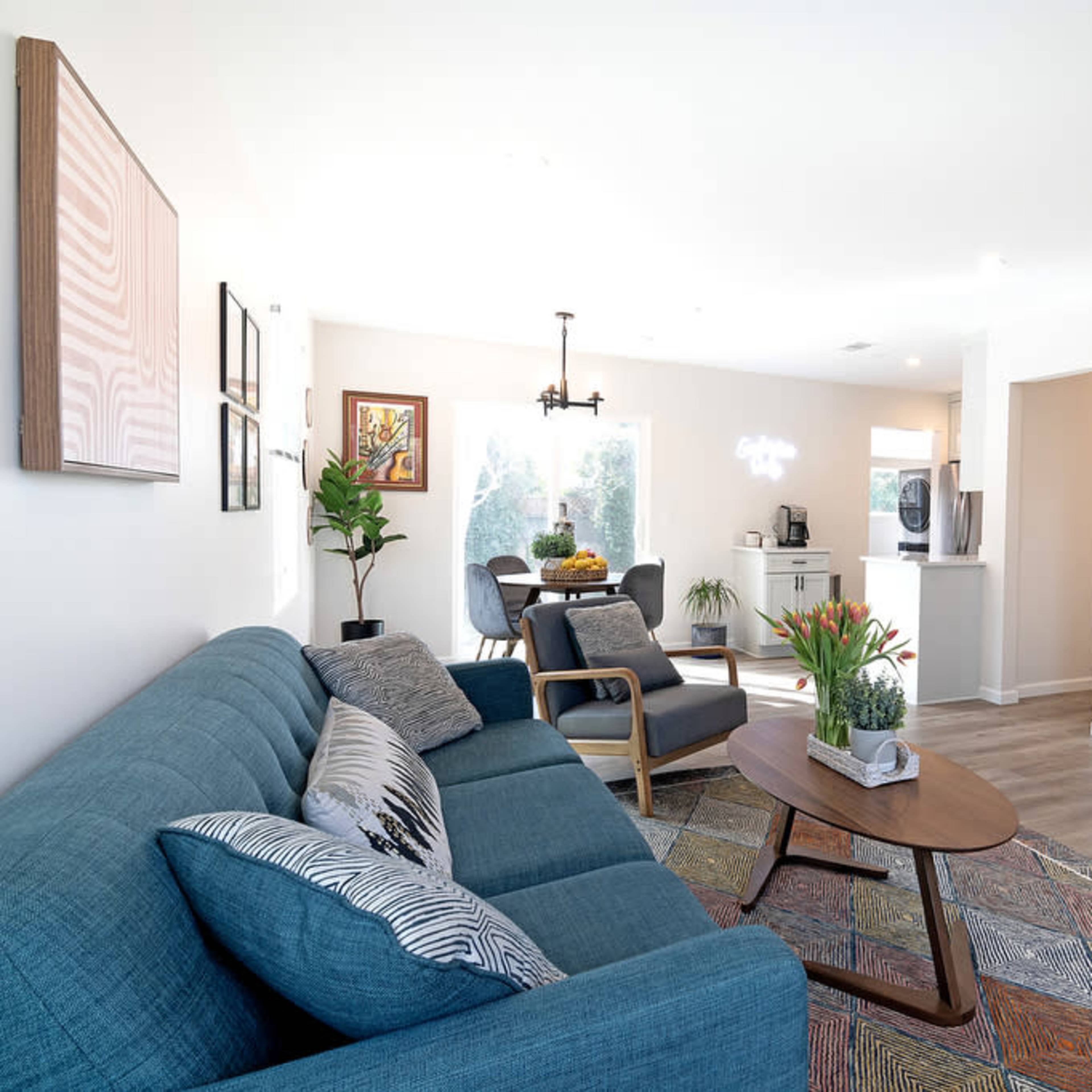 A modern living room with a blue sofa, a round coffee table, and a wall-mounted television, leading to an open kitchen area.