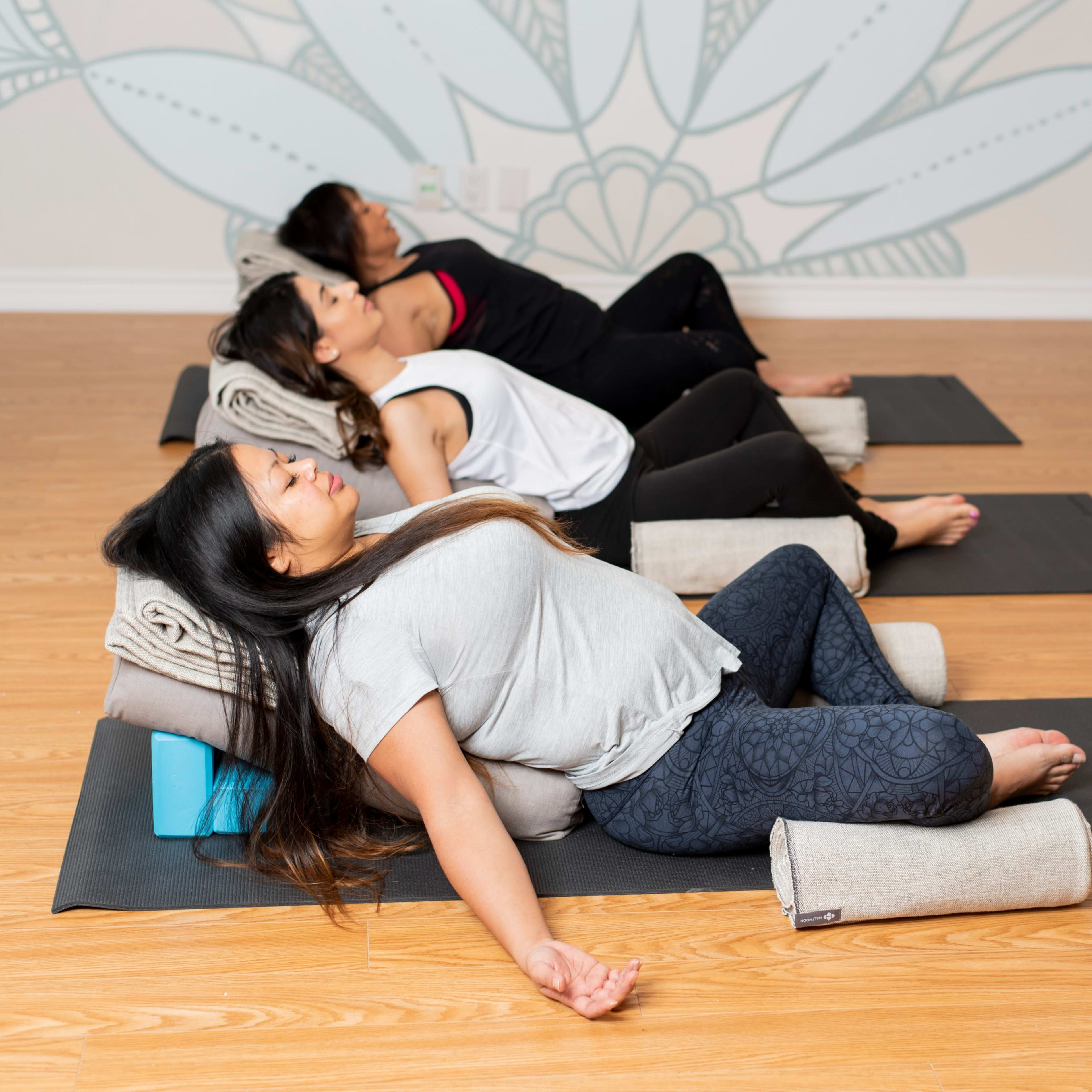 Three women practice restorative yoga on mats in a studio with a decorative wall mural.