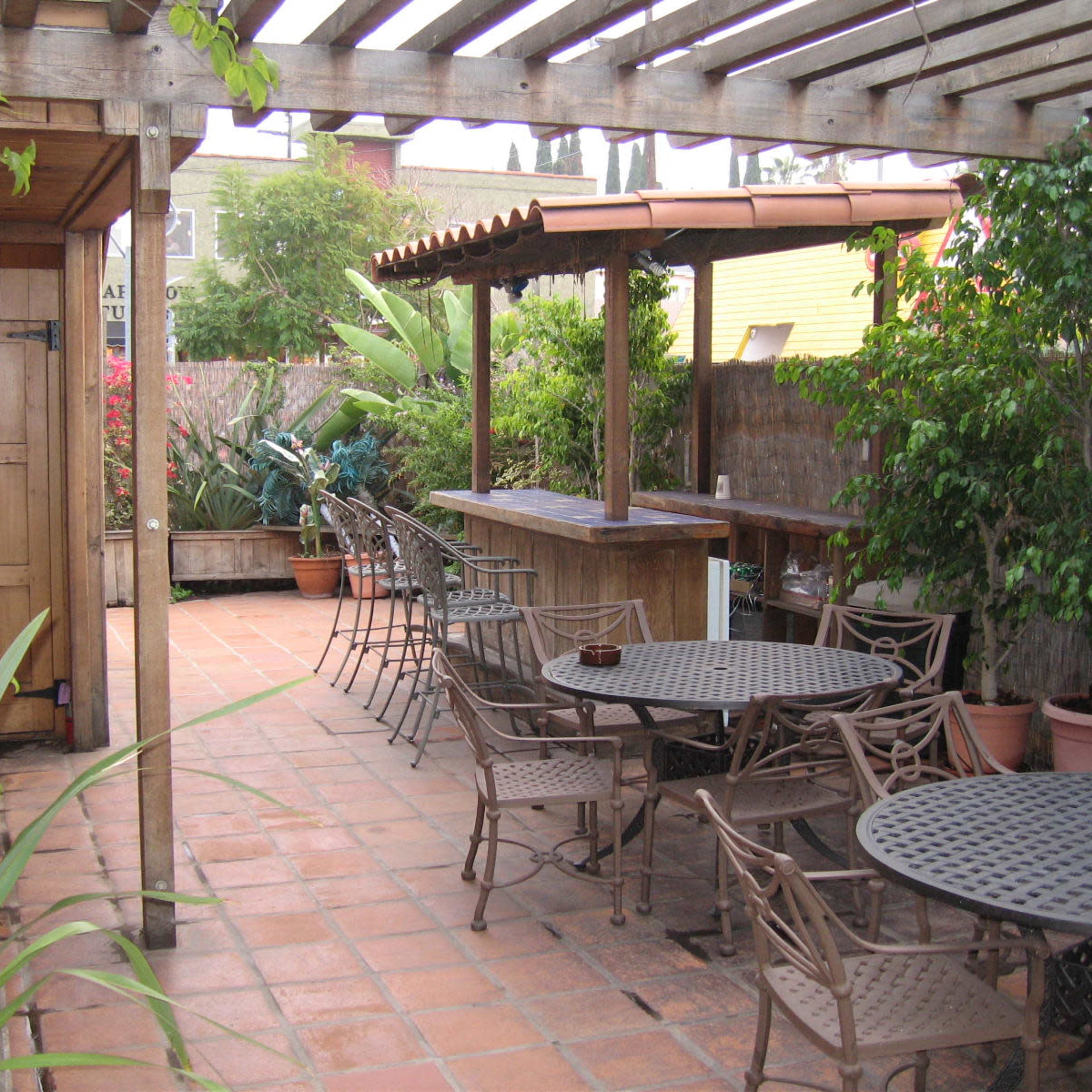 A spacious patio with several metal tables and chairs, surrounded by greenery and featuring a wooden bar under a pergola.
