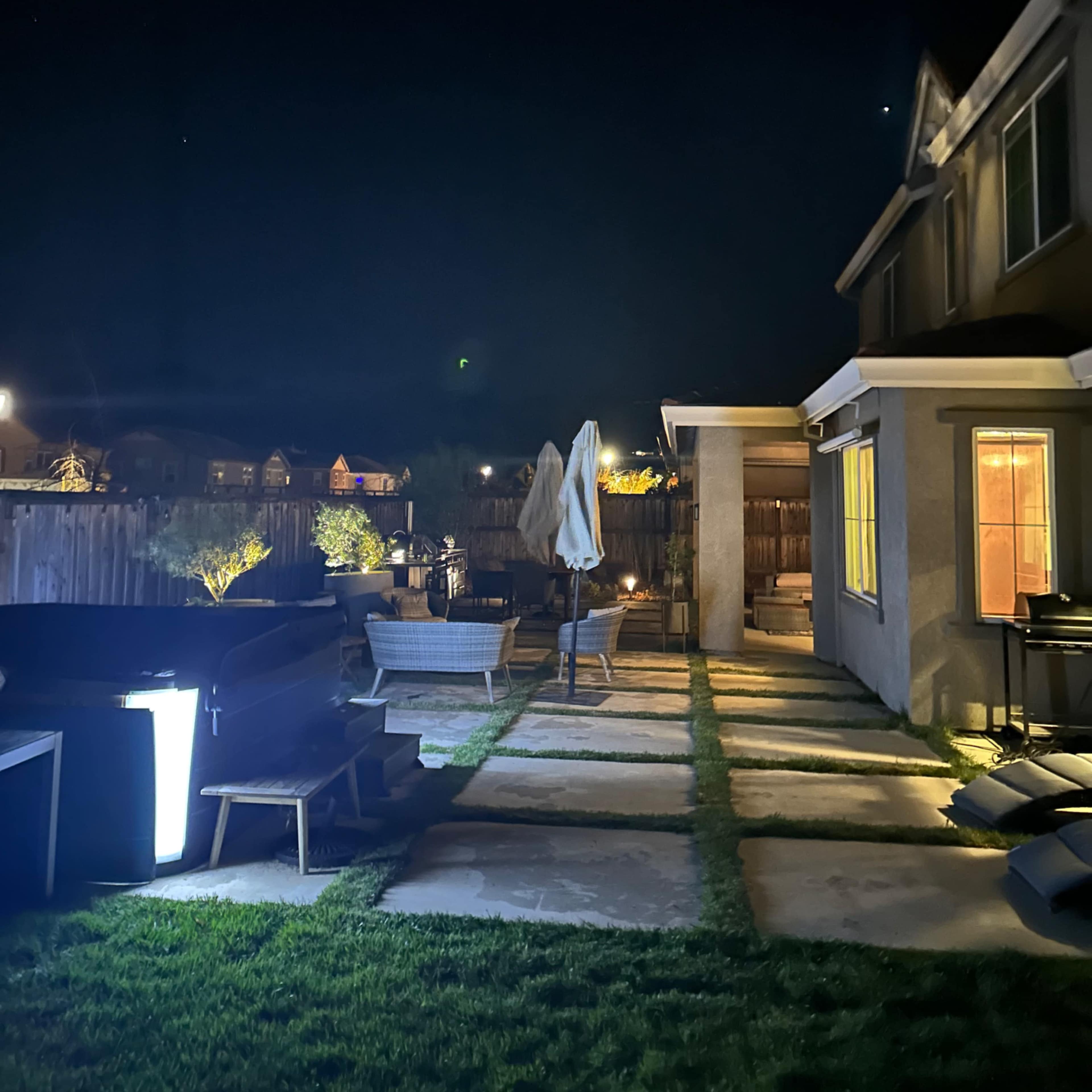 A dimly lit backyard features stone pathways, a patio with chairs, and a house illuminated from the inside at night.