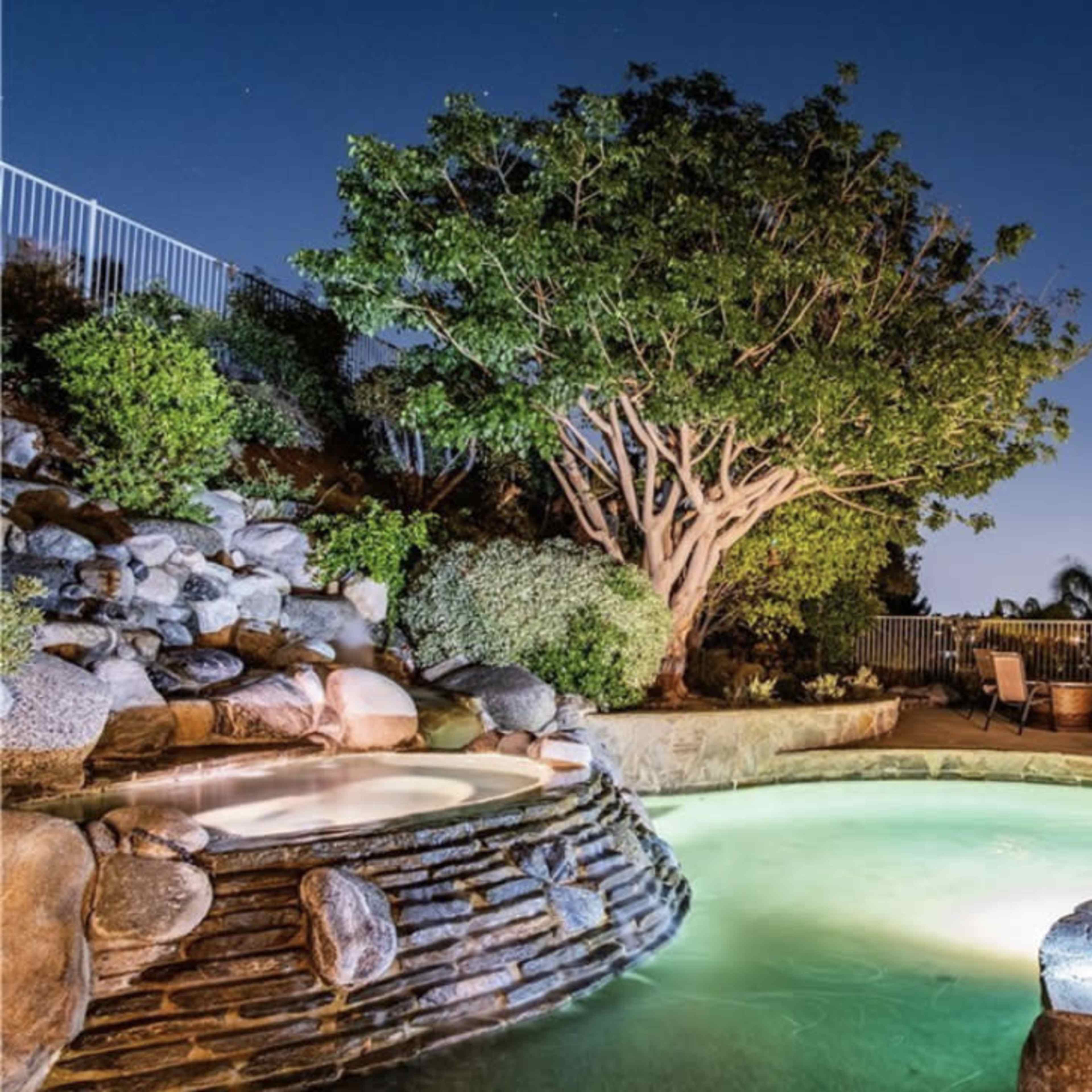 A illuminated backyard features a stone hot tub beside a pool and a large tree at dusk.