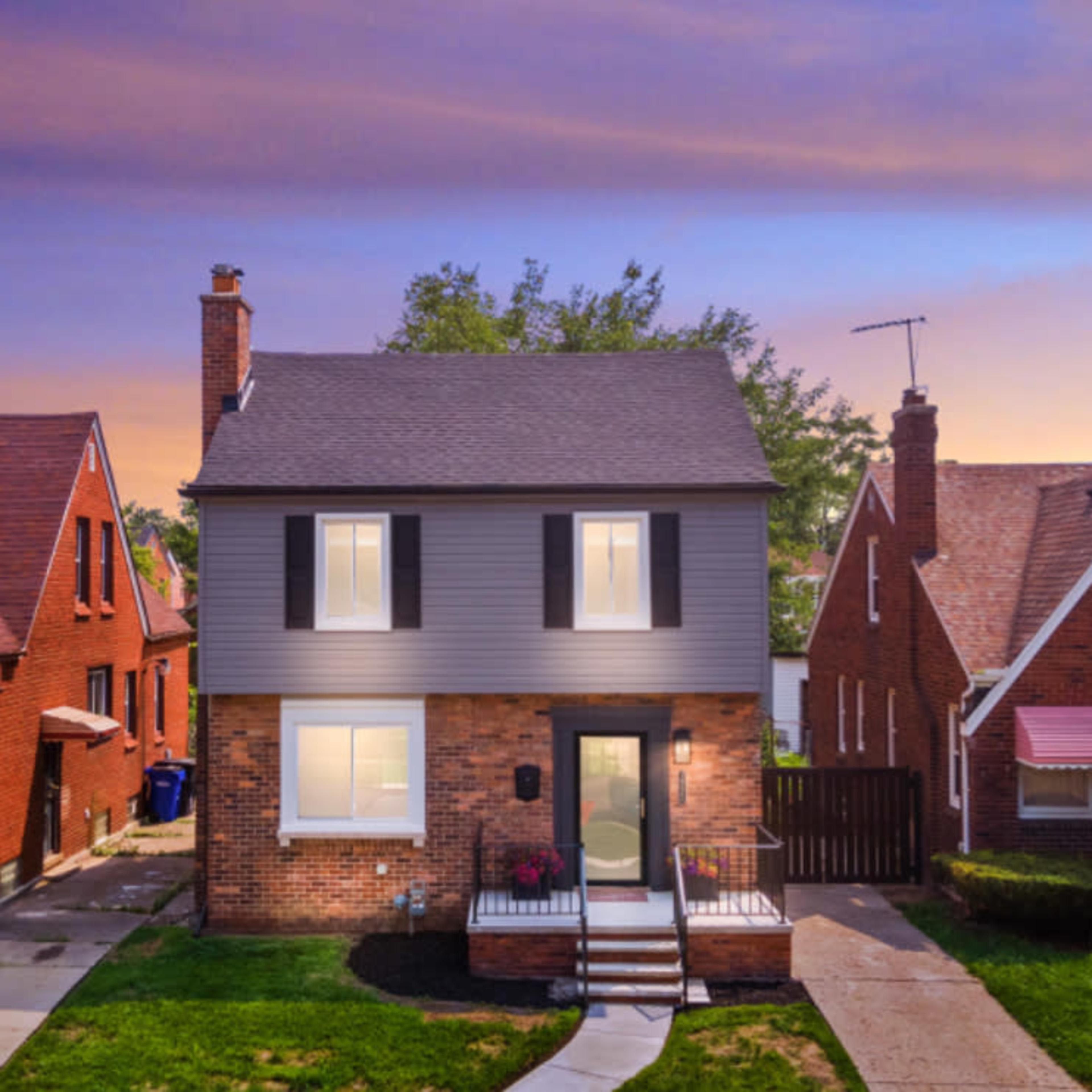 A two-story house with a gray and brick exterior, front porch, and landscaped yard is situated between two other homes at sunset.
