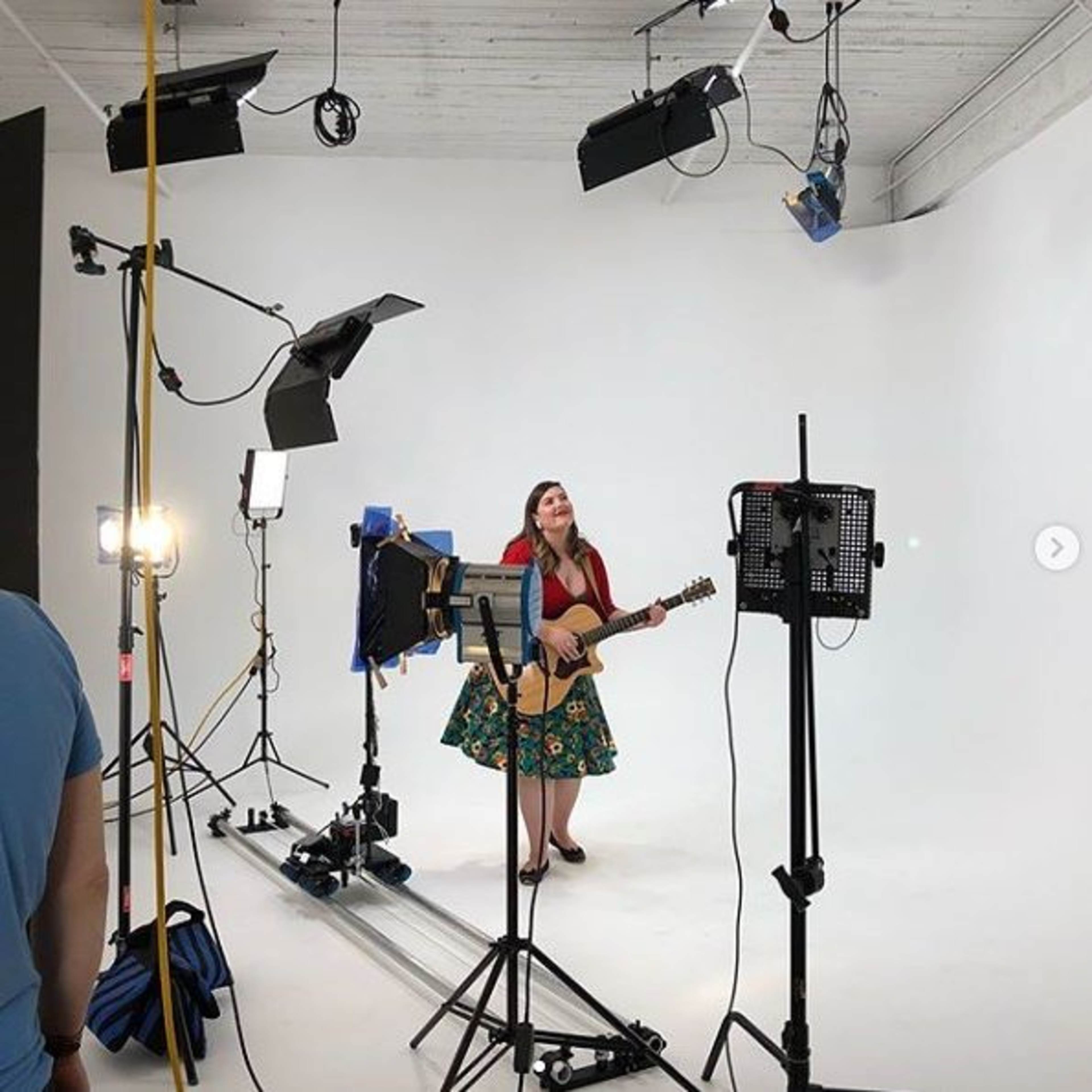 A woman stands in a studio holding a guitar, surrounded by various lighting equipment and cameras on a blank white backdrop.