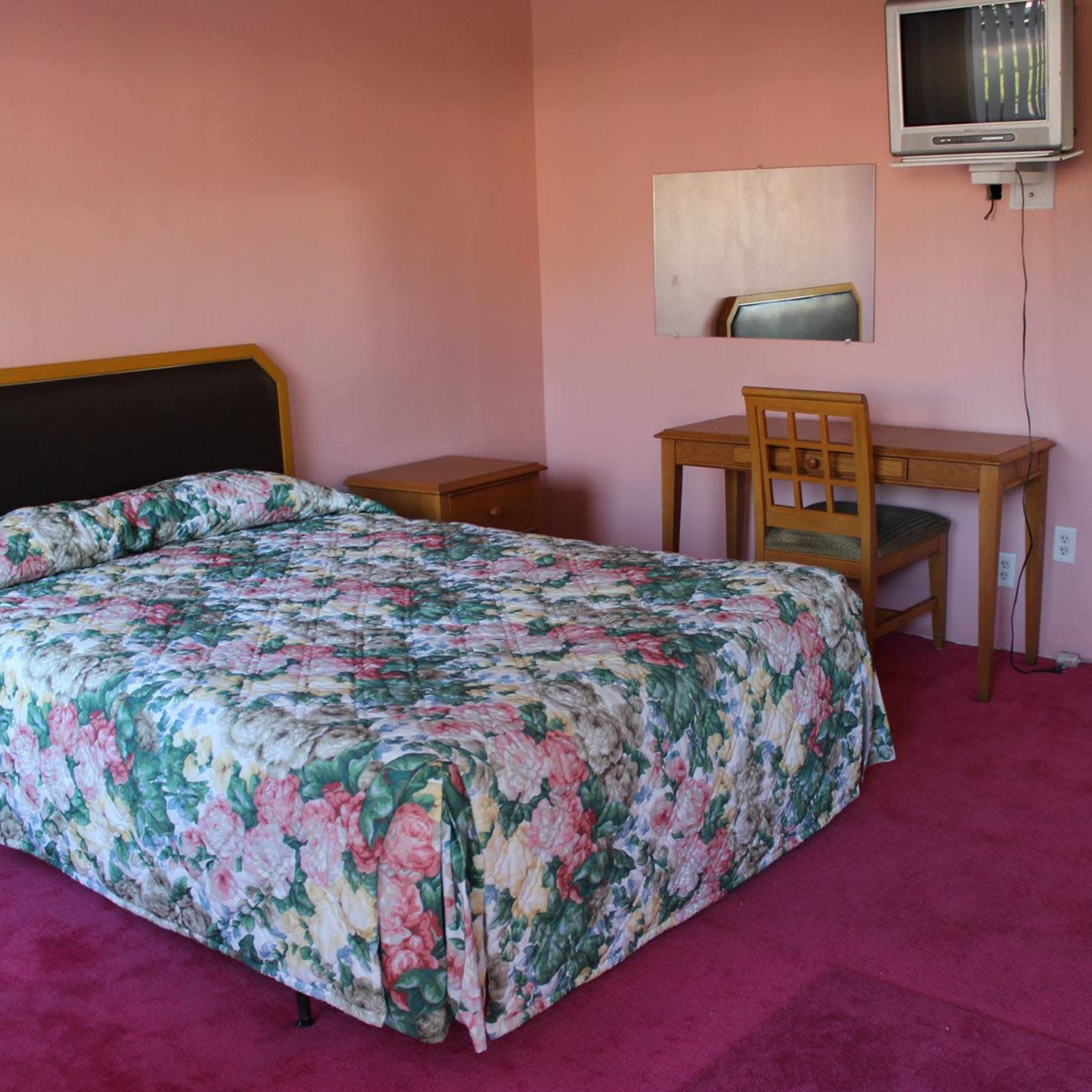 A bedroom with a floral-patterned bedspread, a wooden table with a chair, a small television mounted on the wall, and a window with an air conditioning unit.