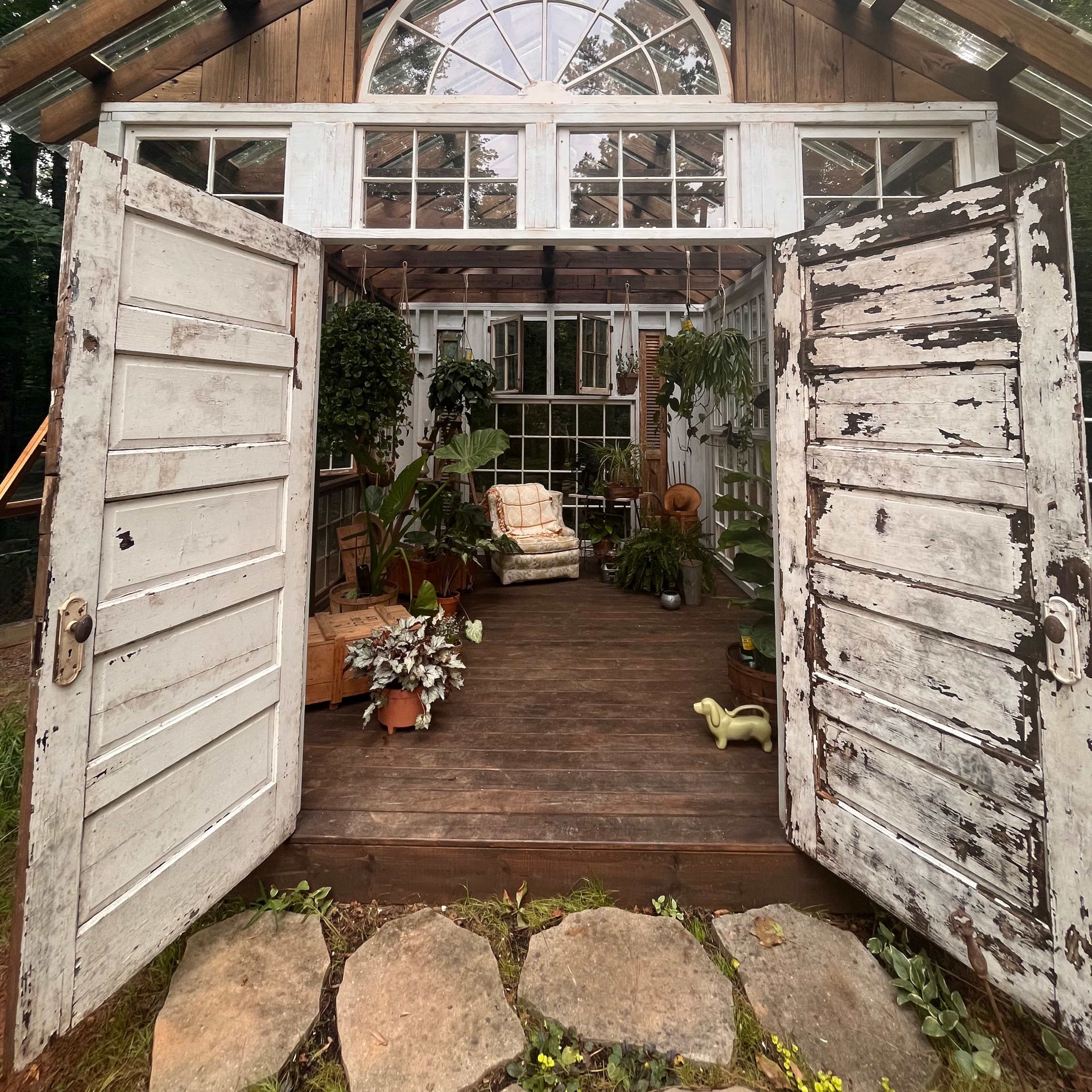 A rustic greenhouse with open double doors showcases a wooden interior adorned with plants and a cozy seating area.