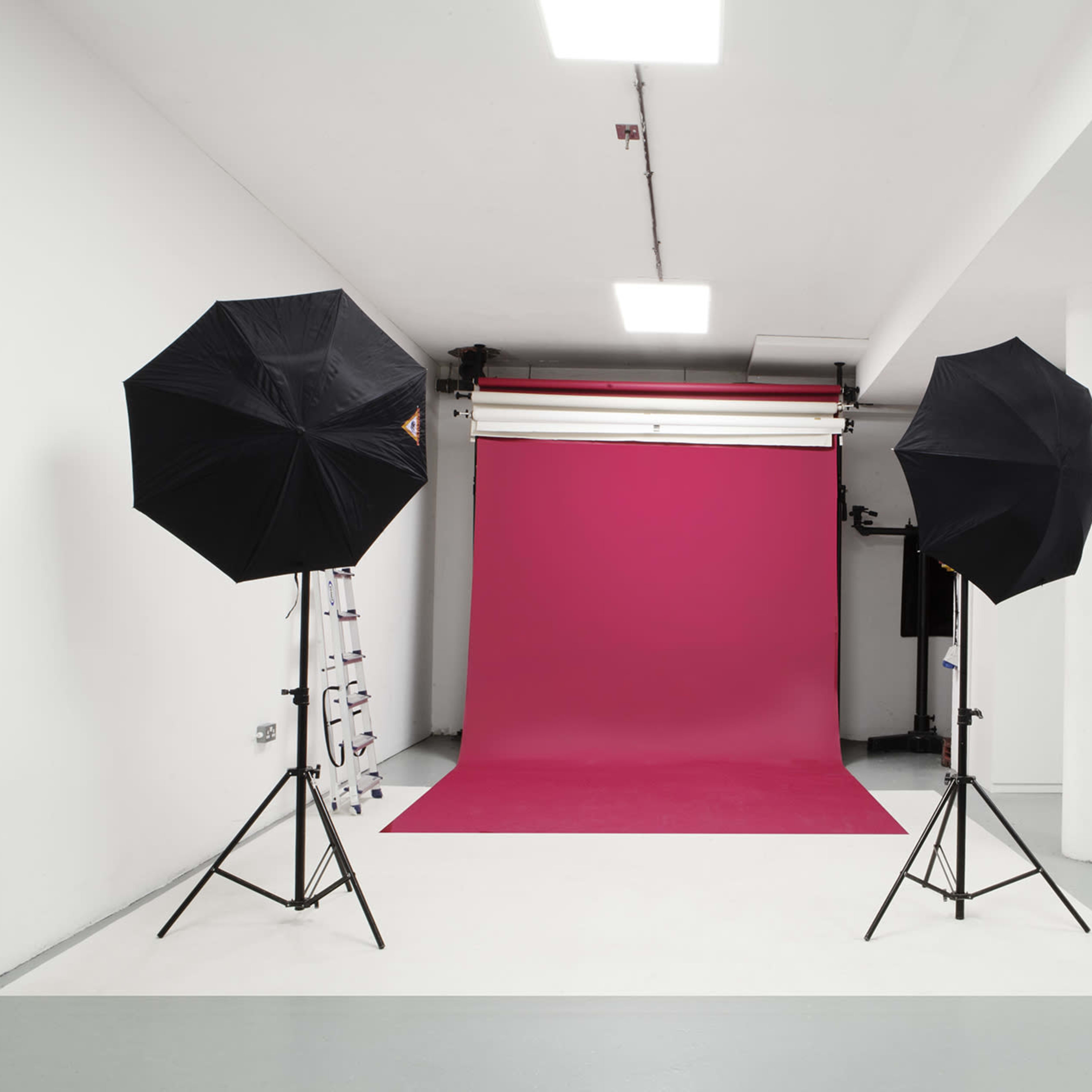 A photography studio with a pink backdrop, two black umbrella light stands, and a white floor.