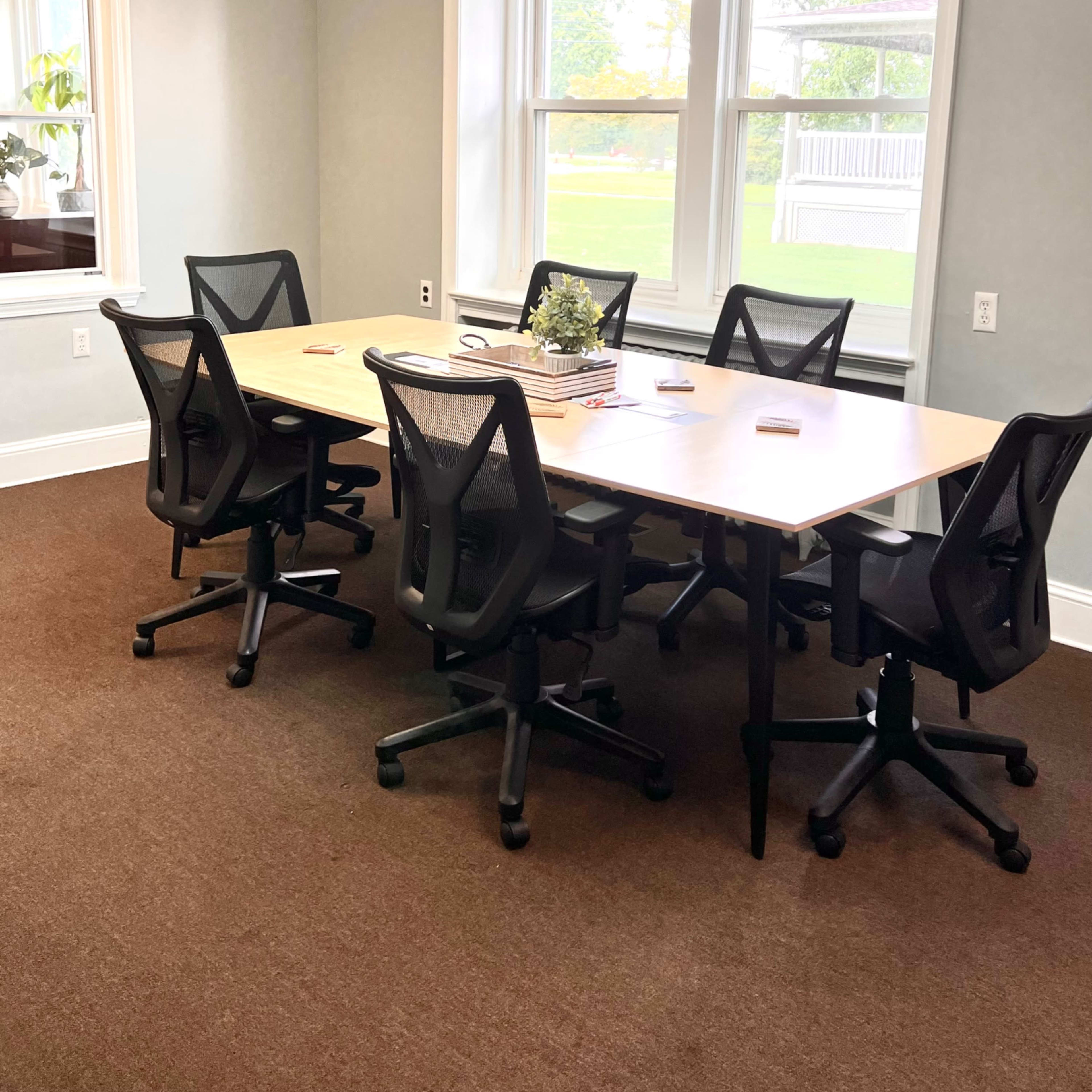 A modern conference room features a large table surrounded by six ergonomic office chairs, with windows providing natural light.