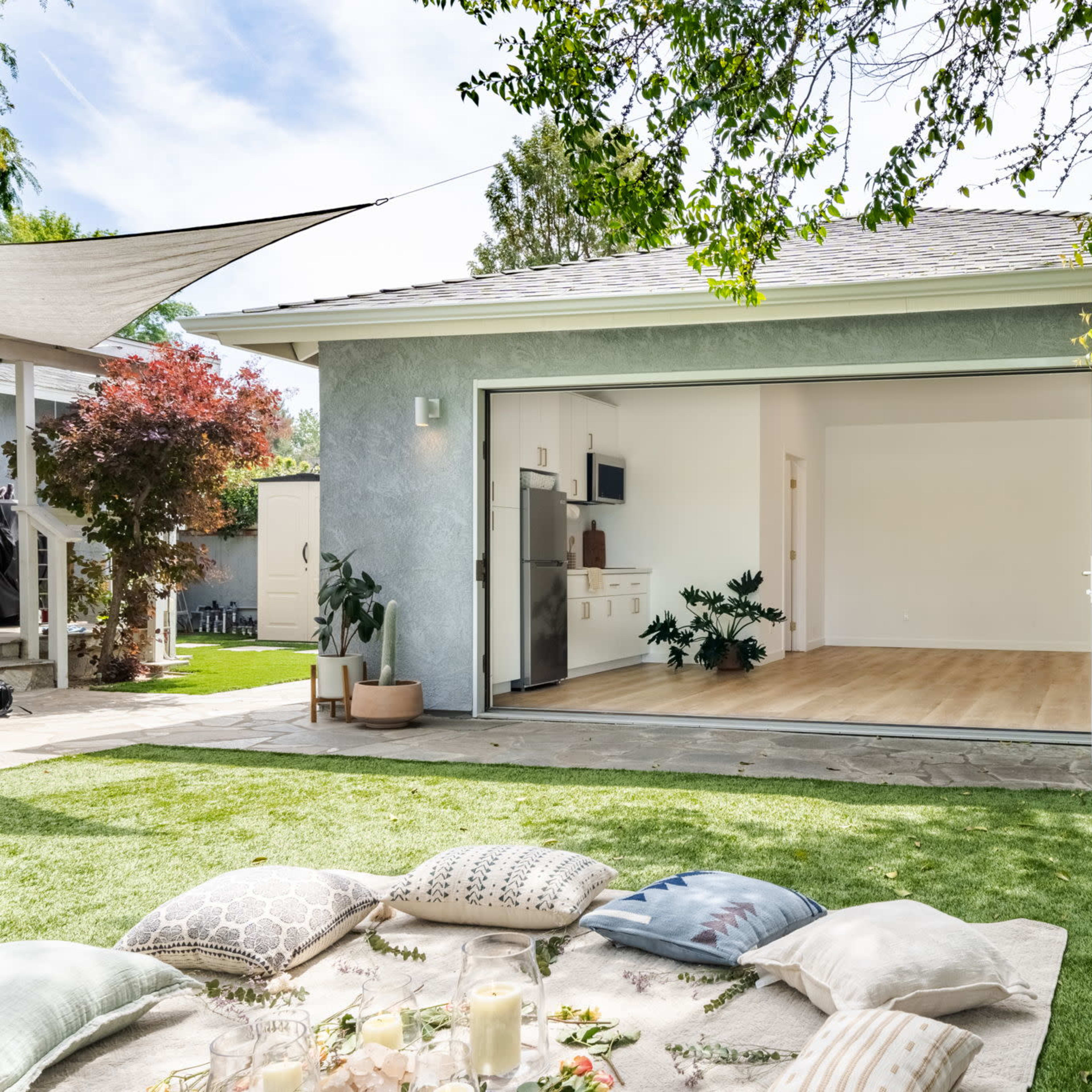 A modern outdoor space features a sliding glass door leading into an open room with wooden flooring, surrounded by greenery and decorative cushions on the grass.