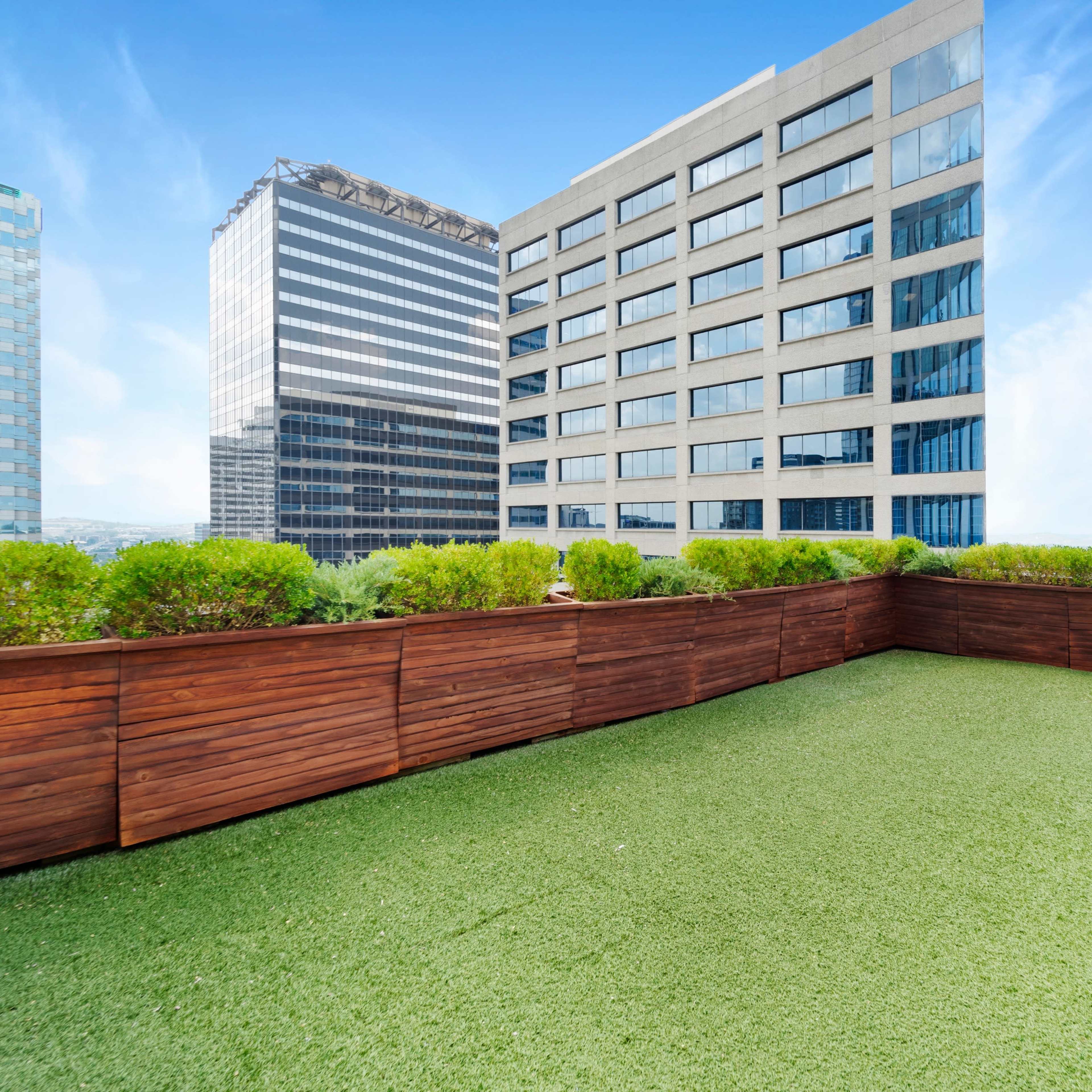 A rooftop terrace with artificial grass and wooden fencing, overlooking adjacent high-rise buildings.