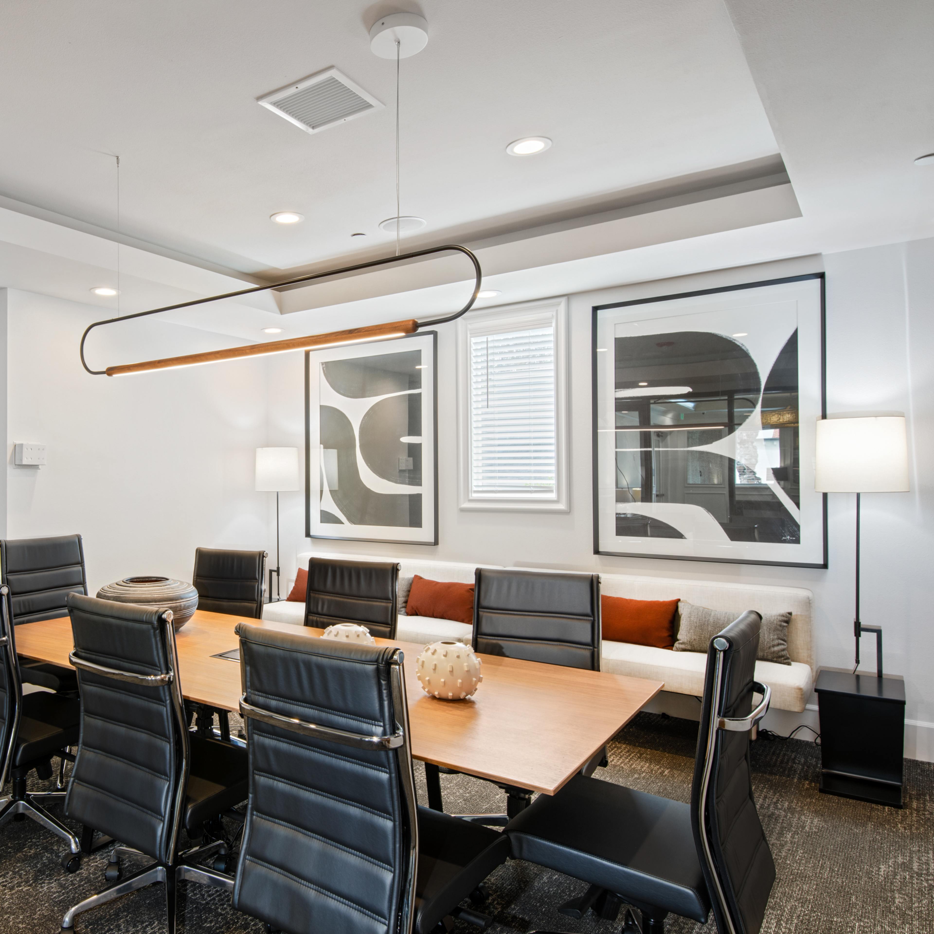 A modern conference room features a long wooden table surrounded by black chairs, with abstract art on the walls and soft seating along one side.