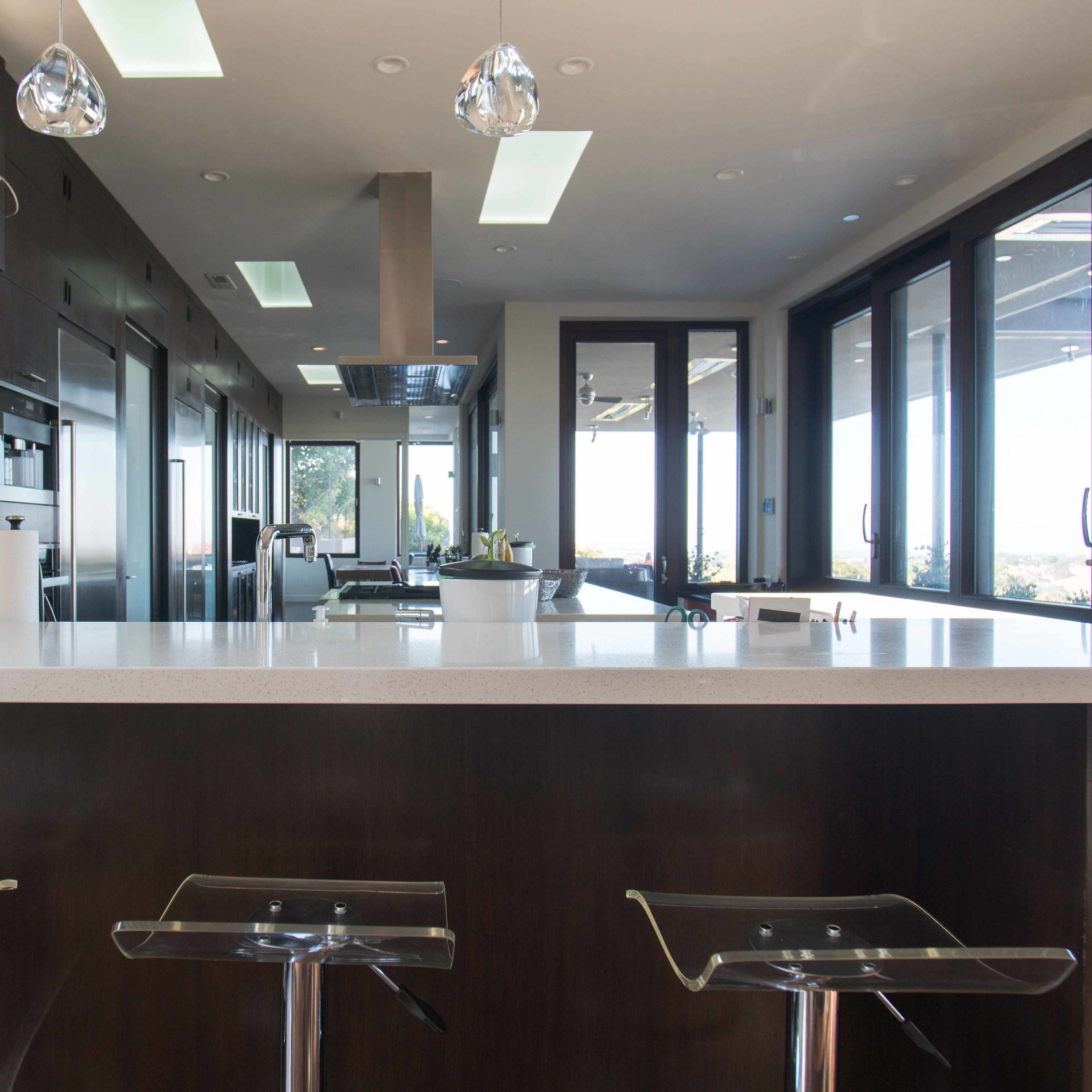 A modern kitchen with a large white countertop, three transparent bar stools, and expansive windows allowing natural light to enter.