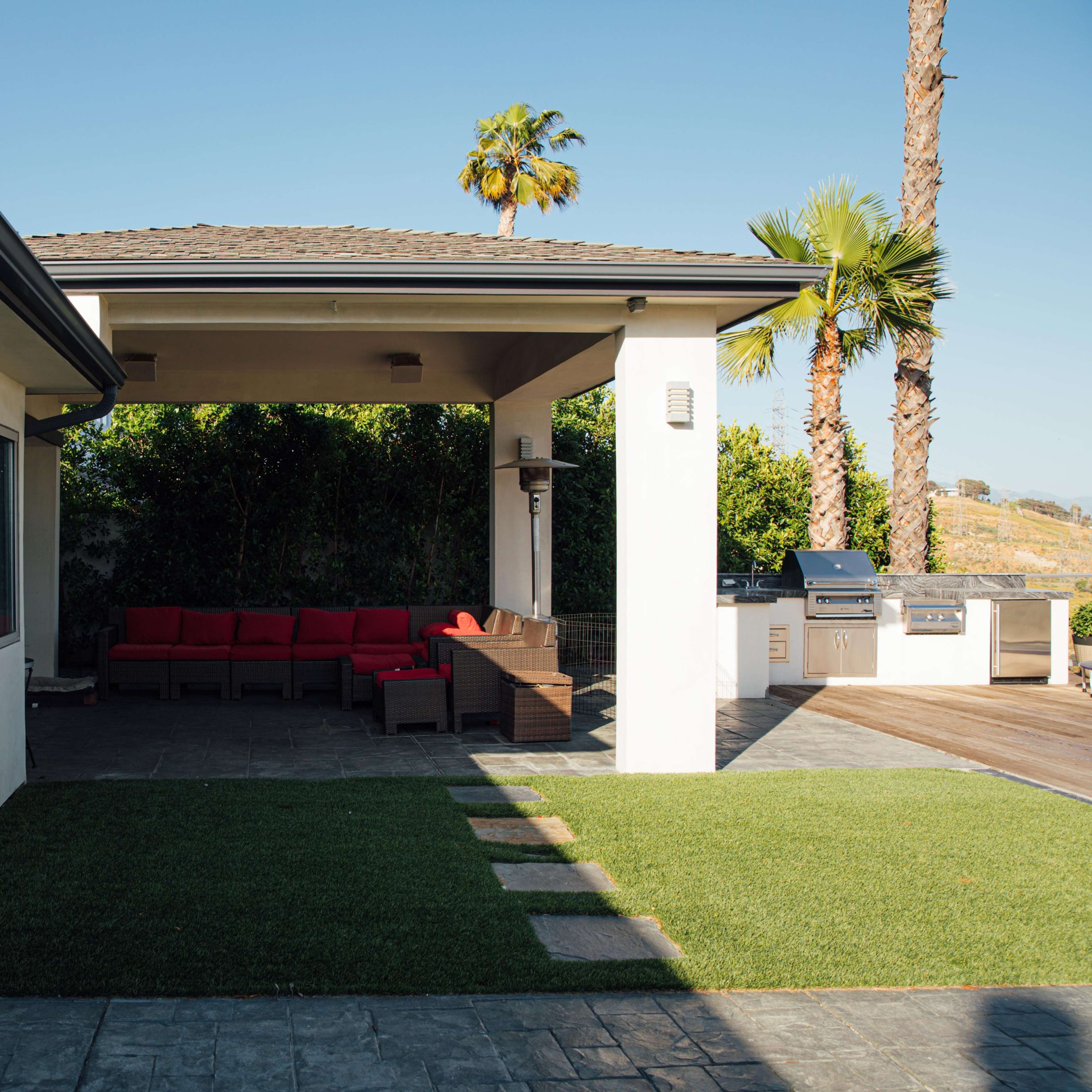 A backyard patio features a seating area with red cushions, a grill, and palm trees in the background.
