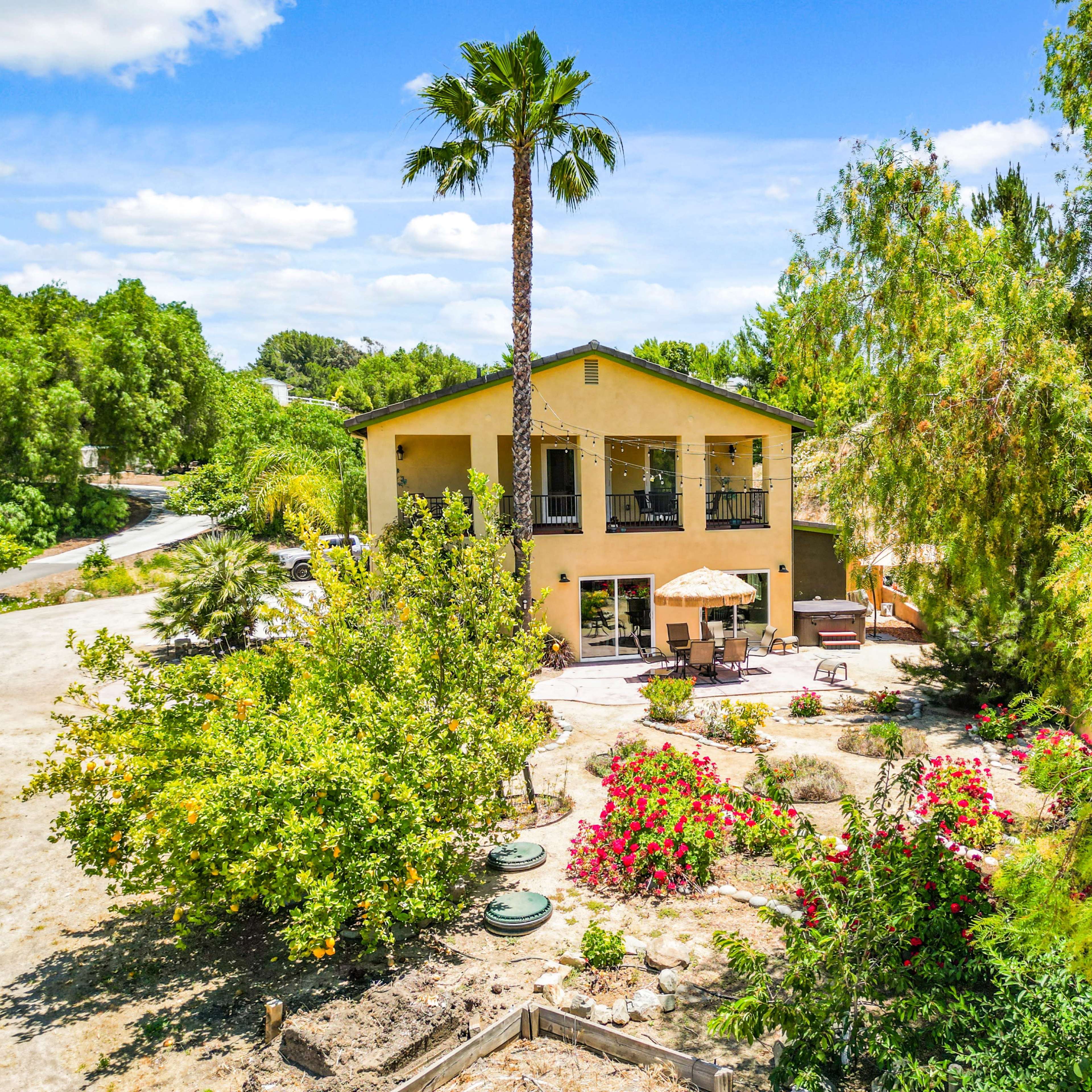 A two-story yellow house with a balcony is surrounded by colorful gardens and palm trees.
