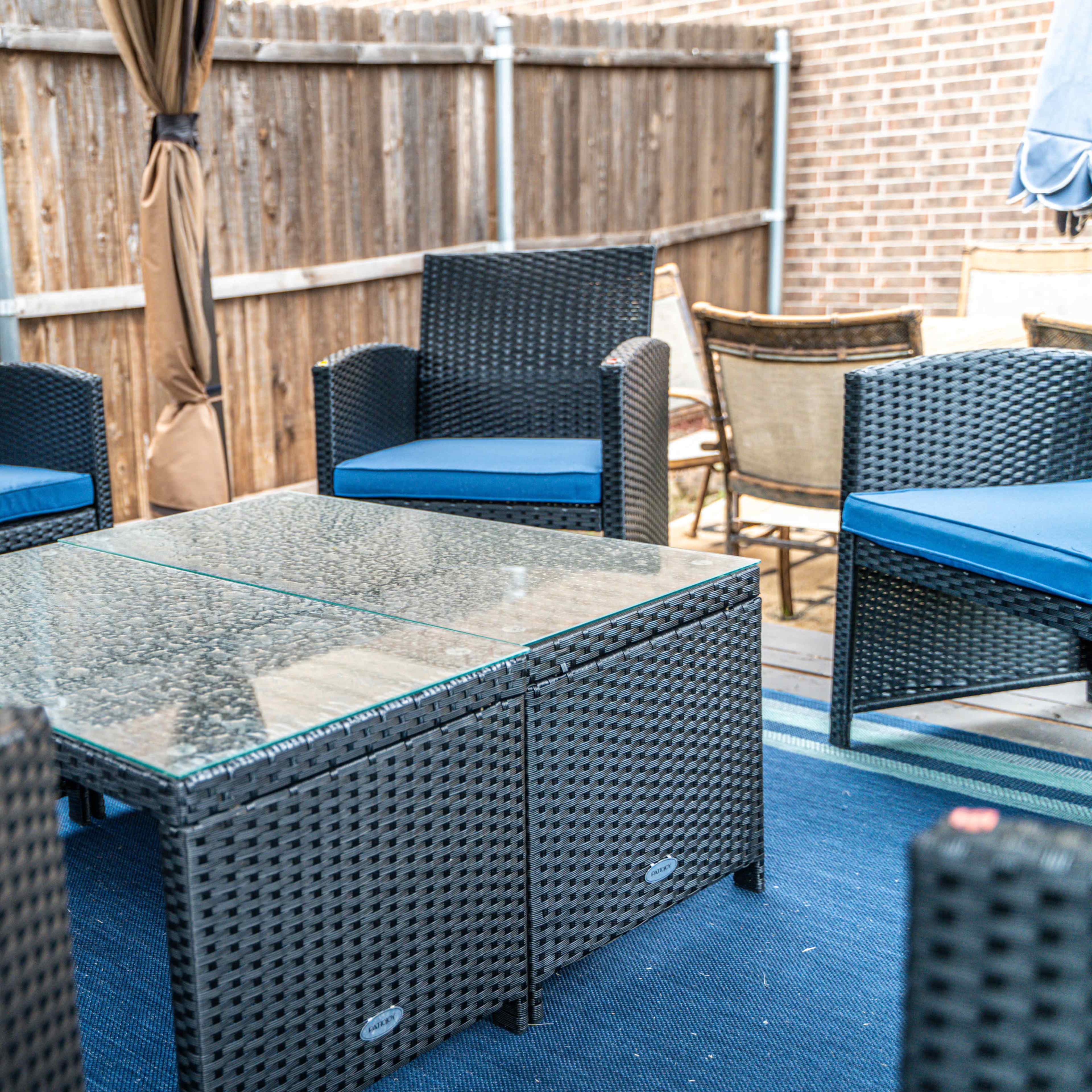 A patio seating arrangement with two wicker sofas, two chairs, and a glass-top coffee table on a blue rug inside a fenced outdoor area.